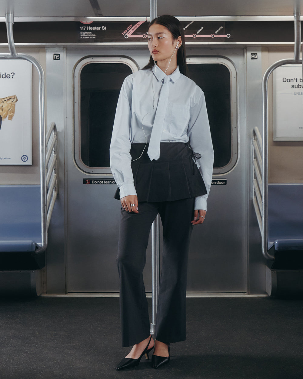 A woman stands in a mostly empty subway car wearing Sundae School FLARE SKIRT PANTS, a light blue button-up shirt with a matching tie, black heels, and earphones. She is looking to the side.
