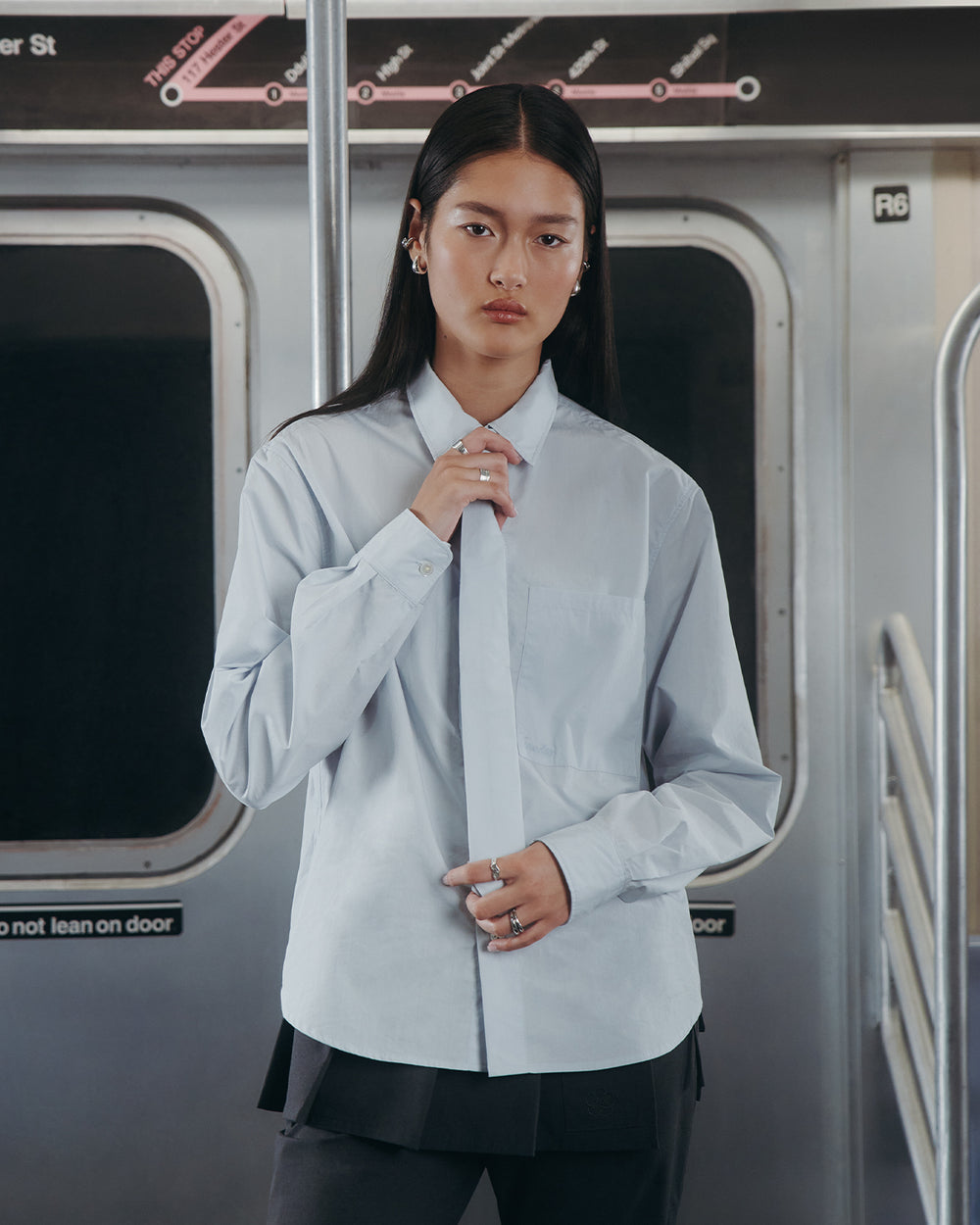 A person with long dark hair wears the Sundae School NECKTIE SHIRT—a relaxed light blue button-up with a removable tie—while standing inside a subway train, with the subway map and window visible in the background.