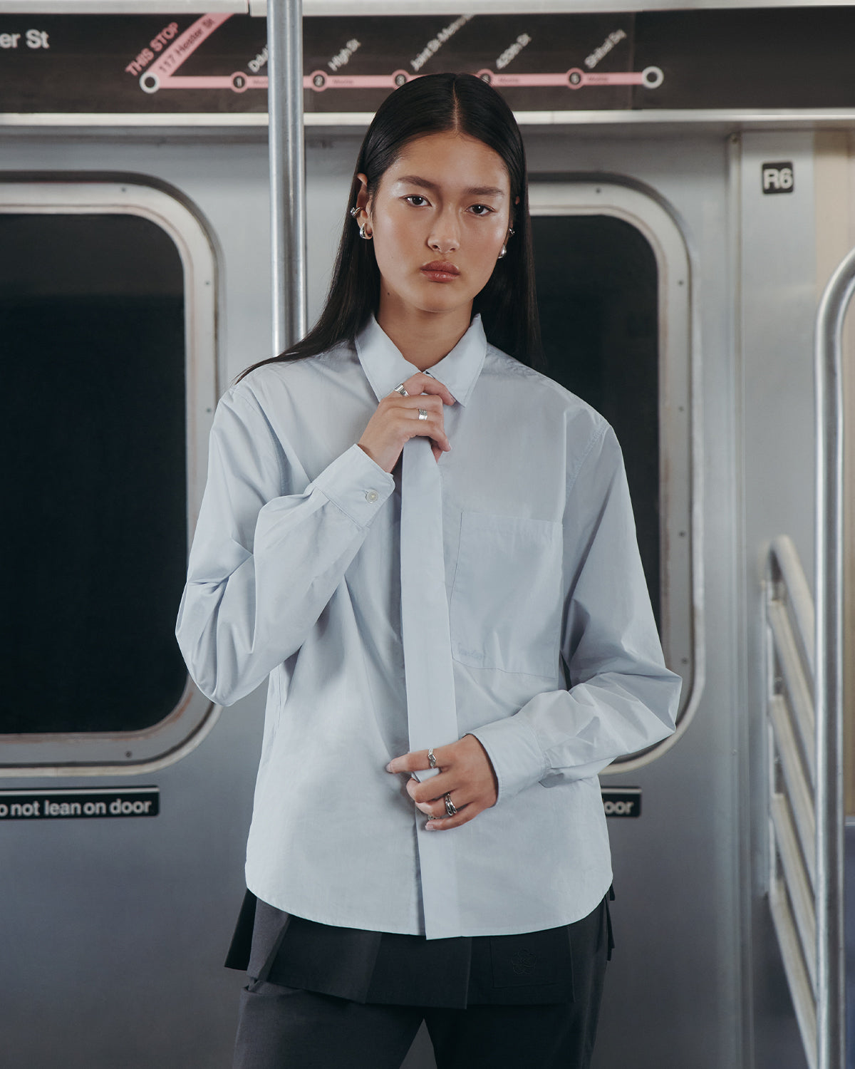 A person with long dark hair wears the Sundae School NECKTIE SHIRT—a relaxed light blue button-up with a removable tie—while standing inside a subway train, with the subway map and window visible in the background.