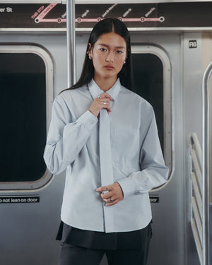 A person with long dark hair wears the Sundae School NECKTIE SHIRT—a relaxed light blue button-up with a removable tie—while standing inside a subway train, with the subway map and window visible in the background.