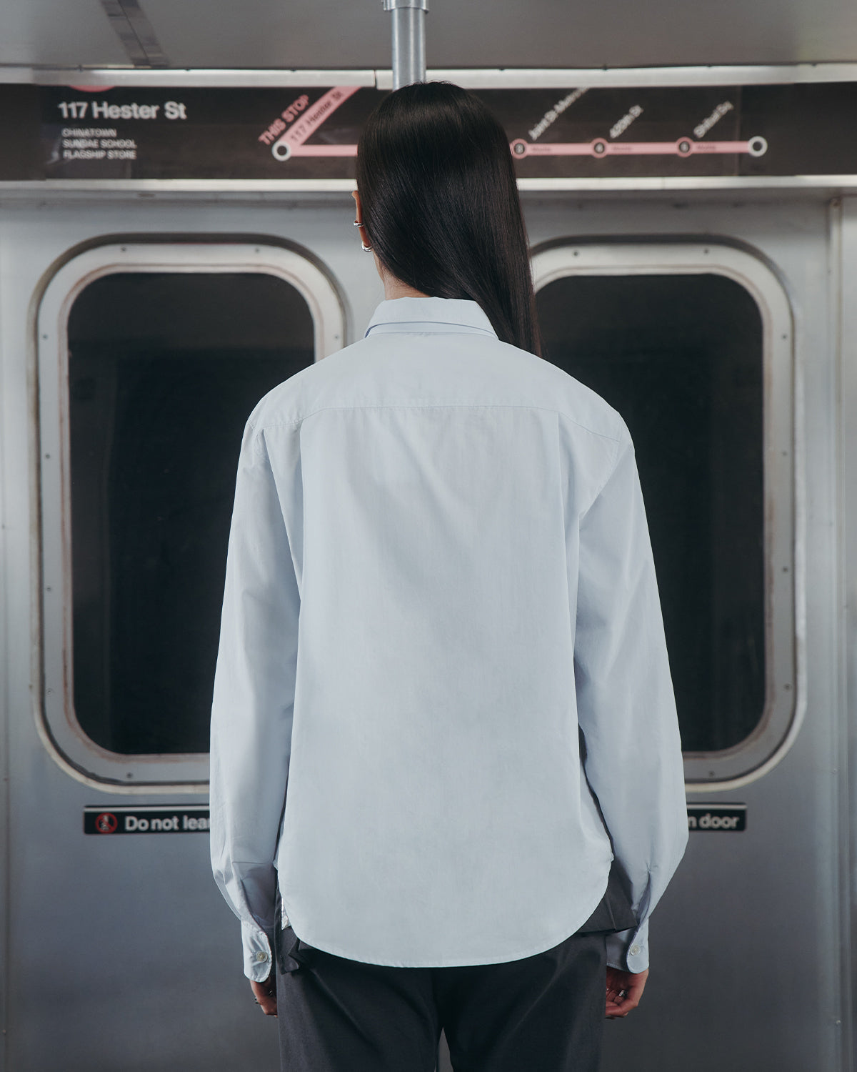 A person with long, straight dark hair wears the Sundae School NECKTIE SHIRT, a relaxed light blue shirt with a stash pocket, standing back to the camera inside a subway at 117 Hester St. A route map is visible above the doors.