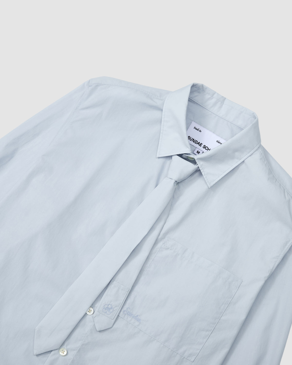 The Sundae School NECKTIE SHIRT features a light blue relaxed fit with a front pocket, visible collar tag, and comes with a matching removable tie, all displayed on a flat white surface.