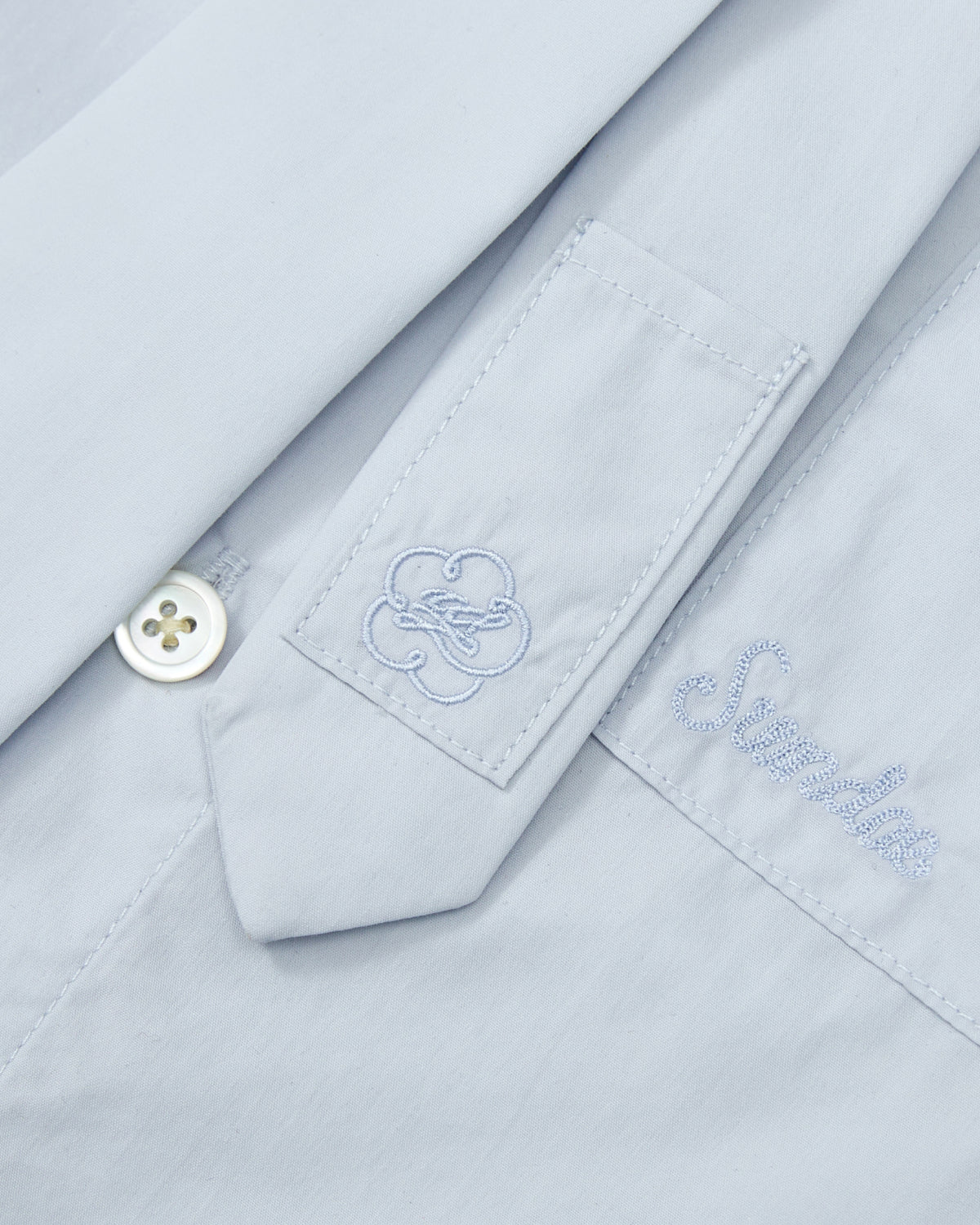 Close-up of the NECKTIE SHIRT by Sundae School, a relaxed fit poplin shirt in light blue, showcasing an embroidered flower-like design on a sleeve tab, a white button, and the word “Smile” stitched above the pocket.