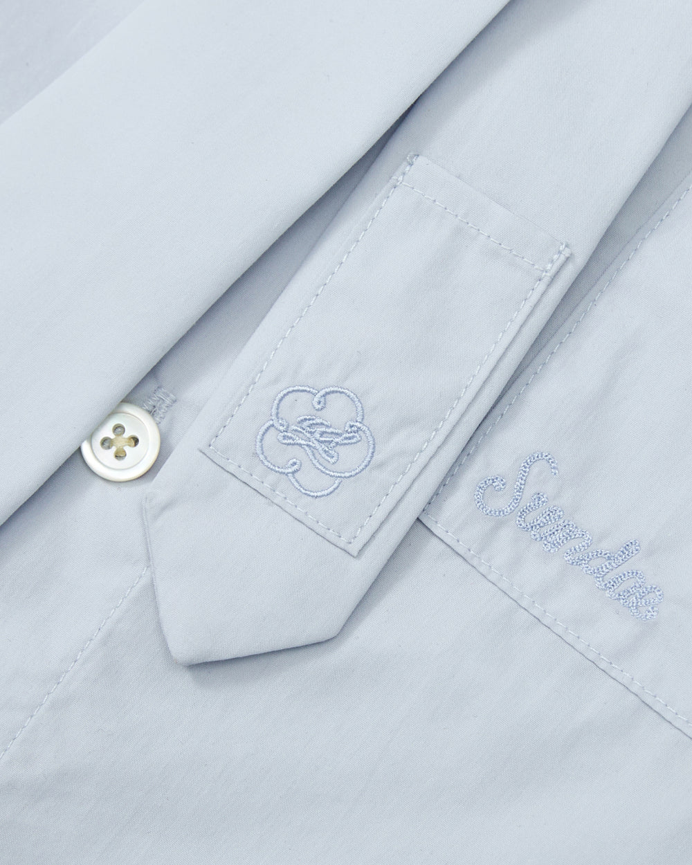 Close-up of the NECKTIE SHIRT by Sundae School, a relaxed fit poplin shirt in light blue, showcasing an embroidered flower-like design on a sleeve tab, a white button, and the word “Smile” stitched above the pocket.