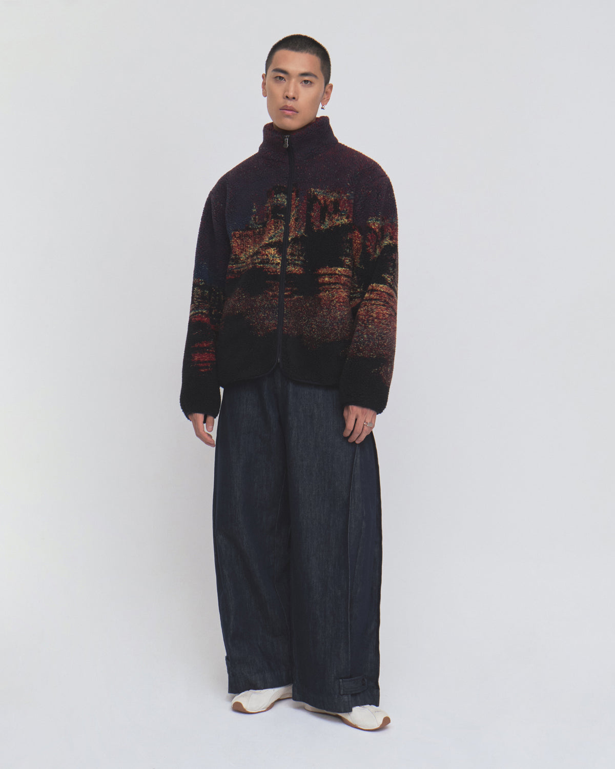 A person stands facing forward, wearing the New Toker Fleece jacket by Sundae School—crafted from dark patterned recycled polyester—with wide-legged dark pants and white shoes against a plain white background.