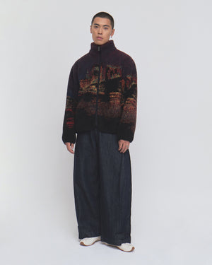 A person stands facing forward, wearing the New Toker Fleece jacket by Sundae School—crafted from dark patterned recycled polyester—with wide-legged dark pants and white shoes against a plain white background.