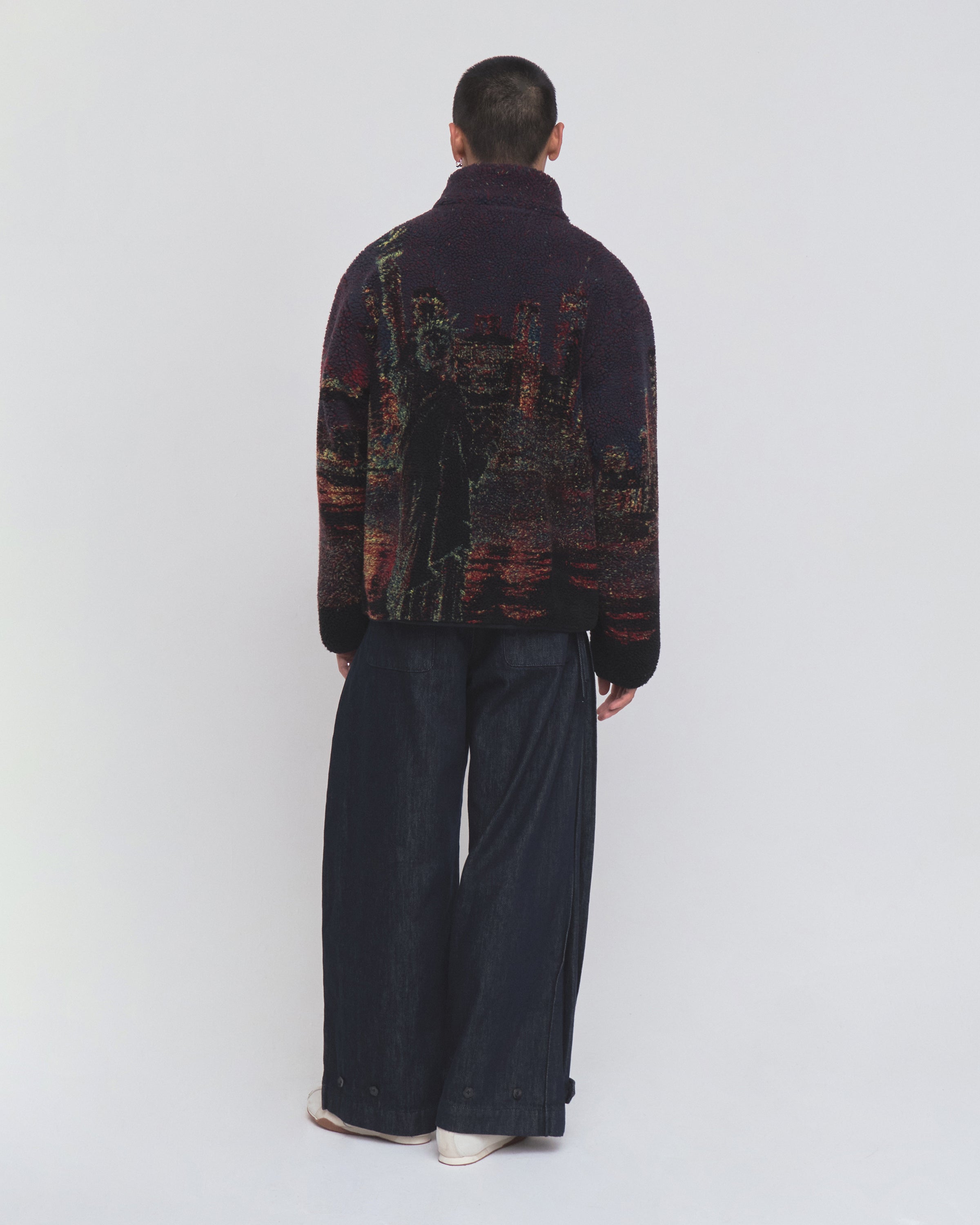 A person stands with their back to the camera, wearing Sundae School’s New Toker Fleece—a dark recycled polyester jacket featuring an image of the Statue of Liberty—paired with wide-leg dark pants, set against a plain light background.