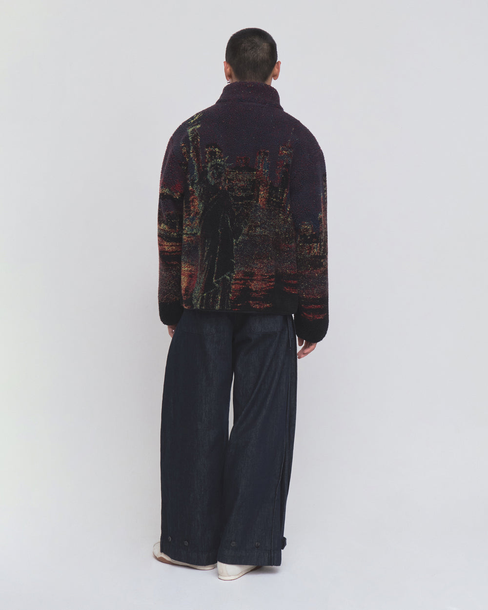 A person stands with their back to the camera, wearing Sundae School’s New Toker Fleece—a dark recycled polyester jacket featuring an image of the Statue of Liberty—paired with wide-leg dark pants, set against a plain light background.
