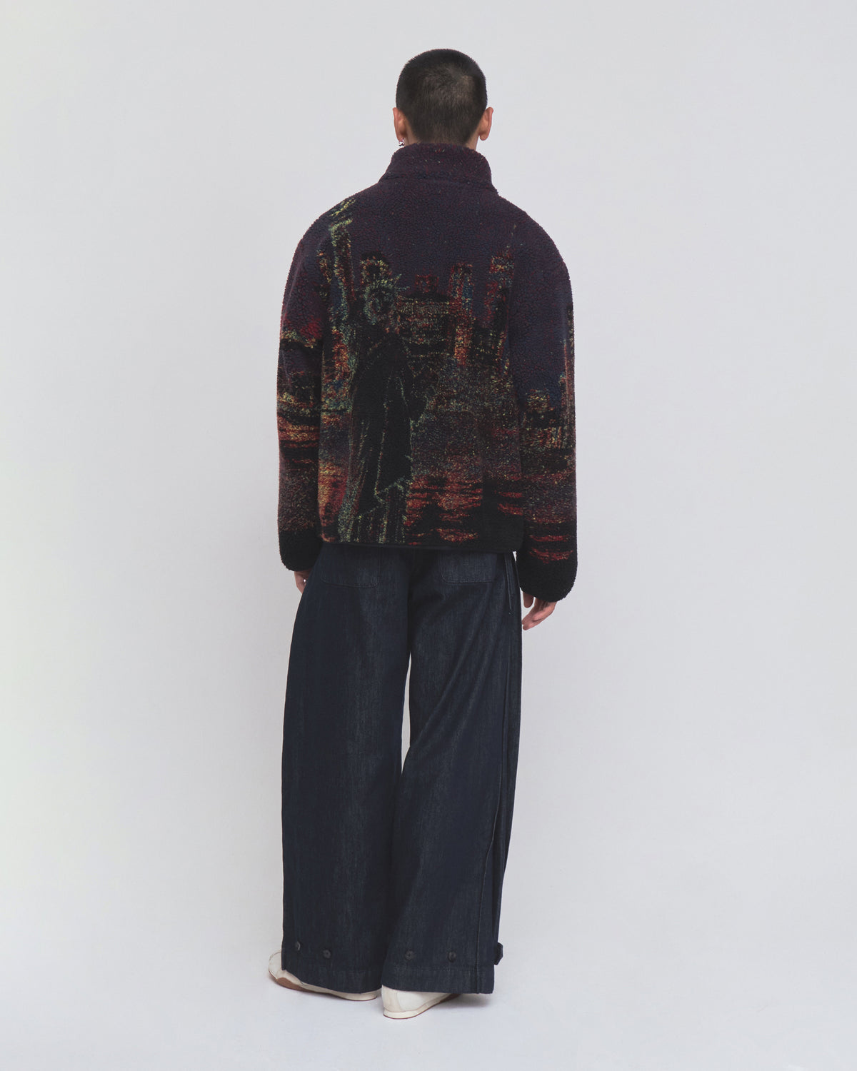 A person stands with their back to the camera, wearing Sundae School’s New Toker Fleece—a dark recycled polyester jacket featuring an image of the Statue of Liberty—paired with wide-leg dark pants, set against a plain light background.