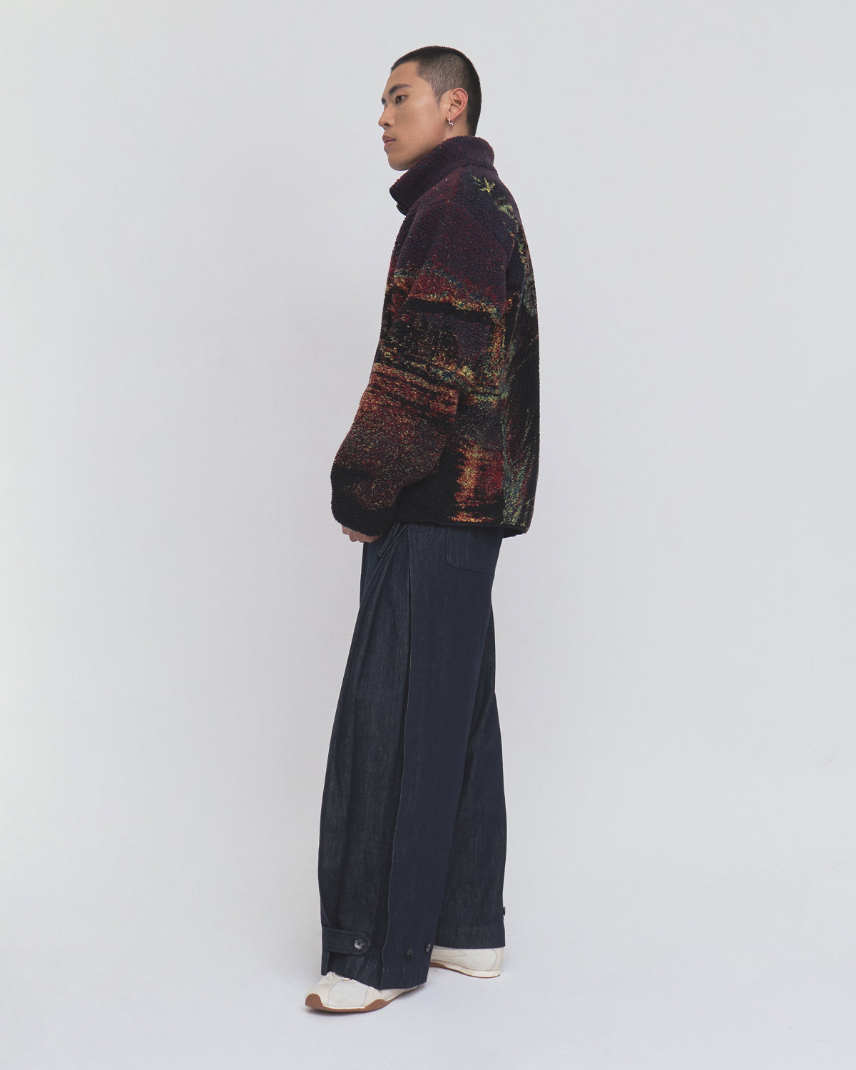 A person stands sideways against a plain white background, wearing Sundae School’s New Toker Fleece—a high-collar jacket crafted from recycled polyester with NYC cityscape jacquard fabric—paired with wide-leg dark blue pants and white shoes.