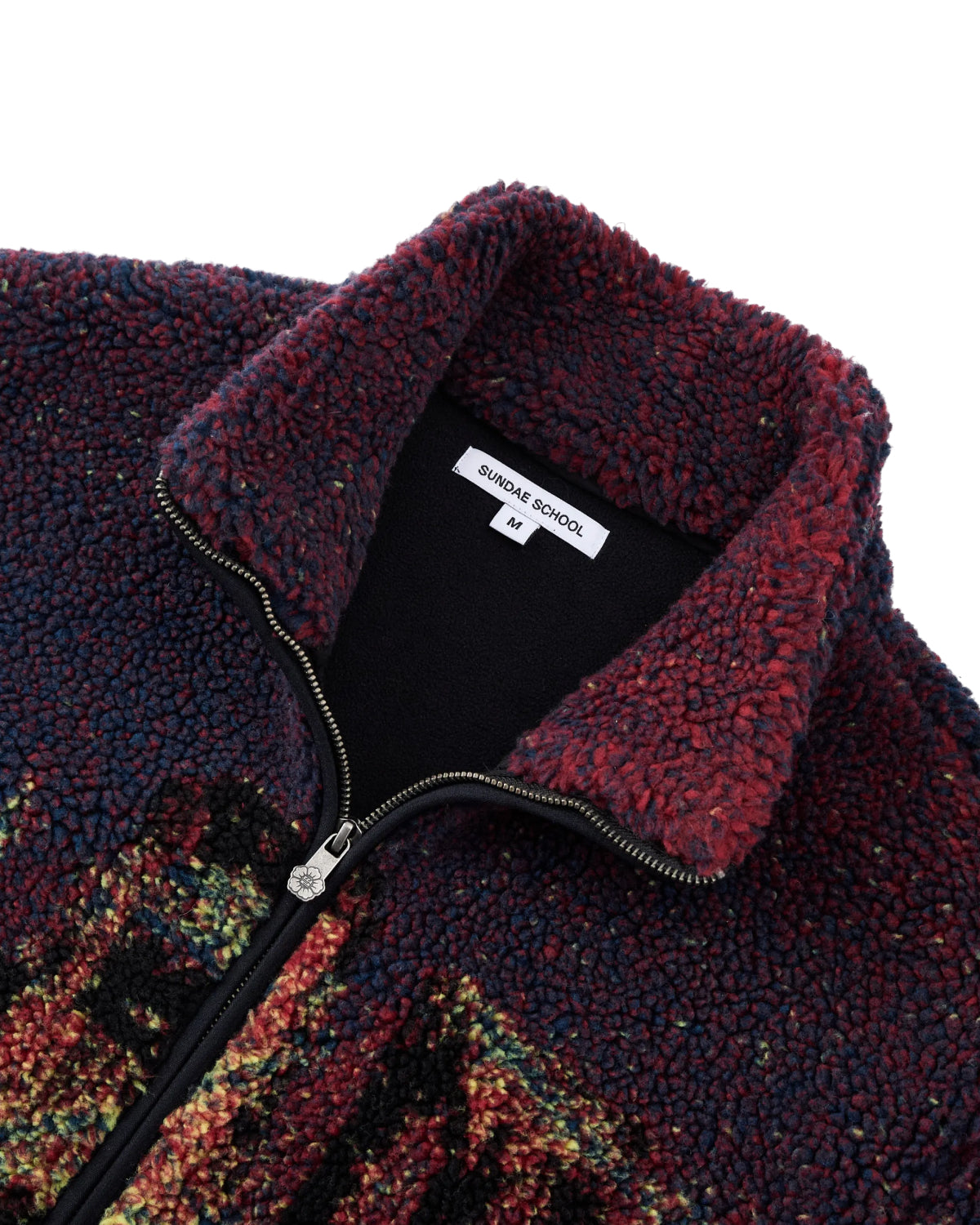 A close-up of the New Toker Fleece by Sundae School, featuring a zippered design in recycled polyester with a textured maroon and navy pattern and a white tag inside the collar that reads Sundae School and M.
