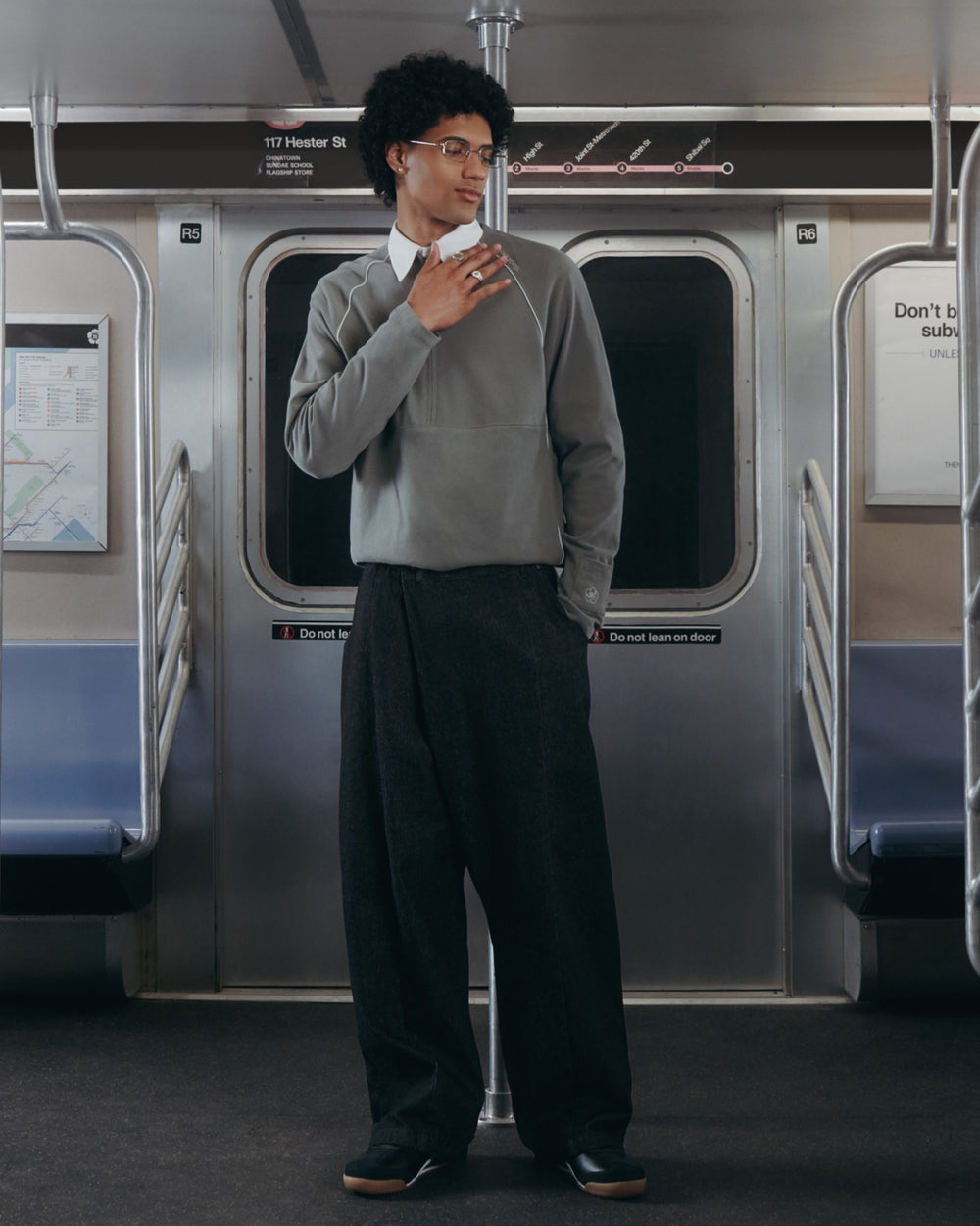 A person with curly hair and glasses wears the Sundae School KHAKI OFFICE QUARTER ZIP over a white shirt and wide-leg dark trousers, standing in an empty subway car, holding a pole and looking sideways with visible subway signs.