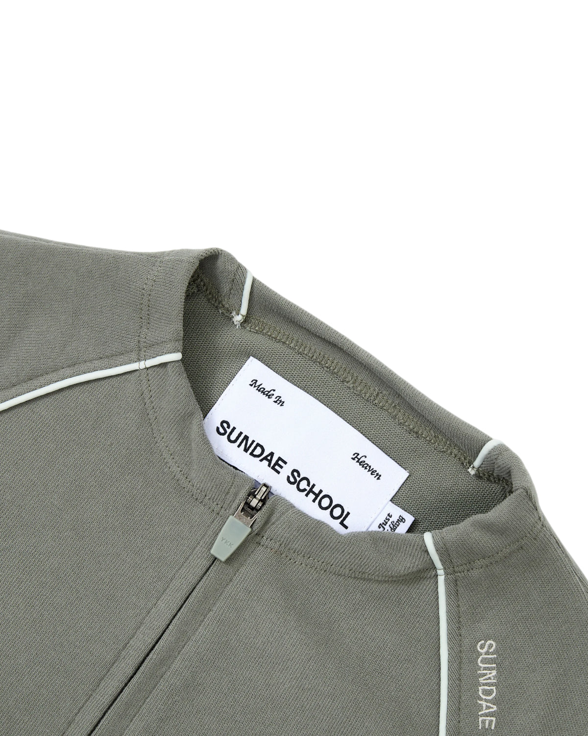 Close-up of the KHAKI OFFICE QUARTER ZIP by Sundae School, crafted from khaki-green cotton jersey with white piping detail. The inner label displays Sundae School and the text “Made In Heaven.” “SUNDAE” is printed near the collar, and a discreet stash pocket enhances the functionality of this quarter-zip design.