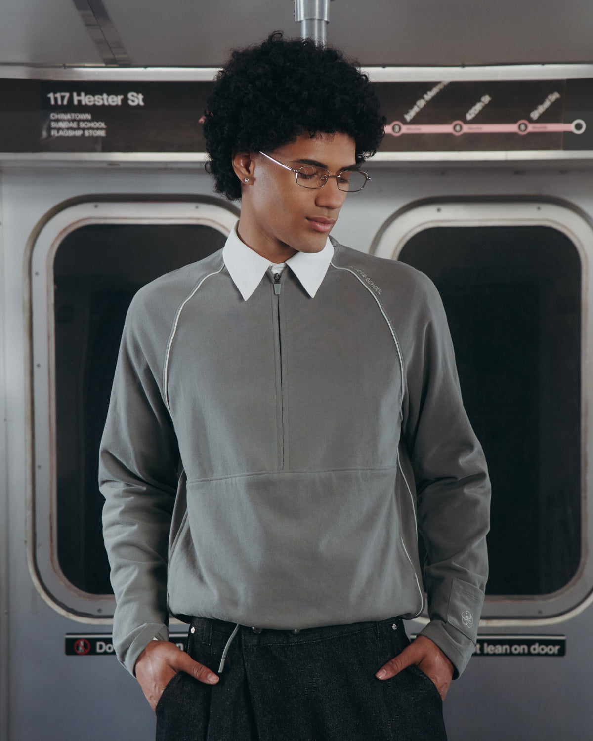 A person with curly hair and glasses wears a Sundae School KHAKI OFFICE QUARTER ZIP in a subway car, hands in pockets, with a subway map and 