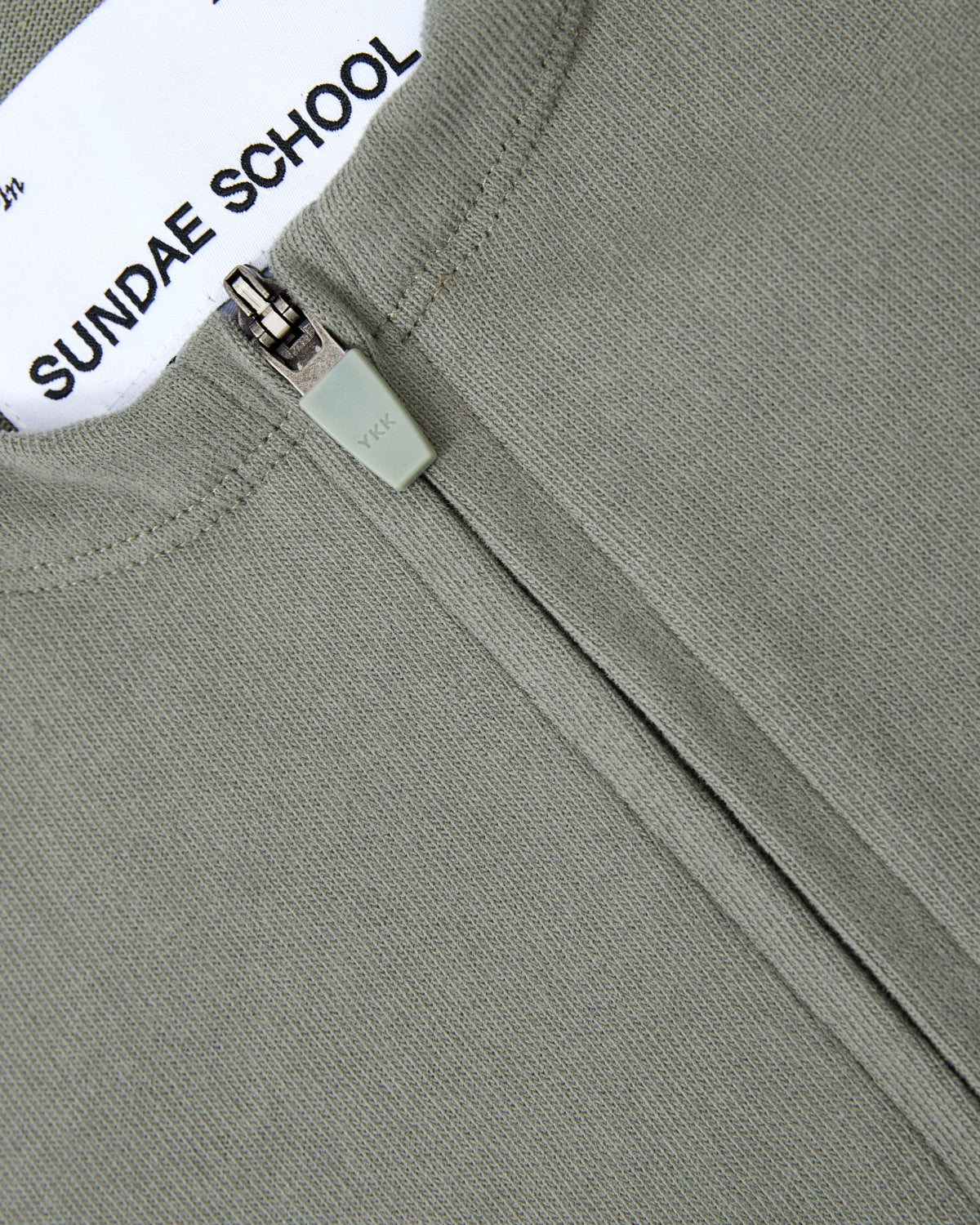 Close-up of the KHAKI OFFICE QUARTER ZIP by Sundae School, featuring a grey cotton jersey knit fabric and a white label with the brand’s name. The partially closed zipper draws attention to its soft material, while a discreet stash pocket may offer additional functionality.