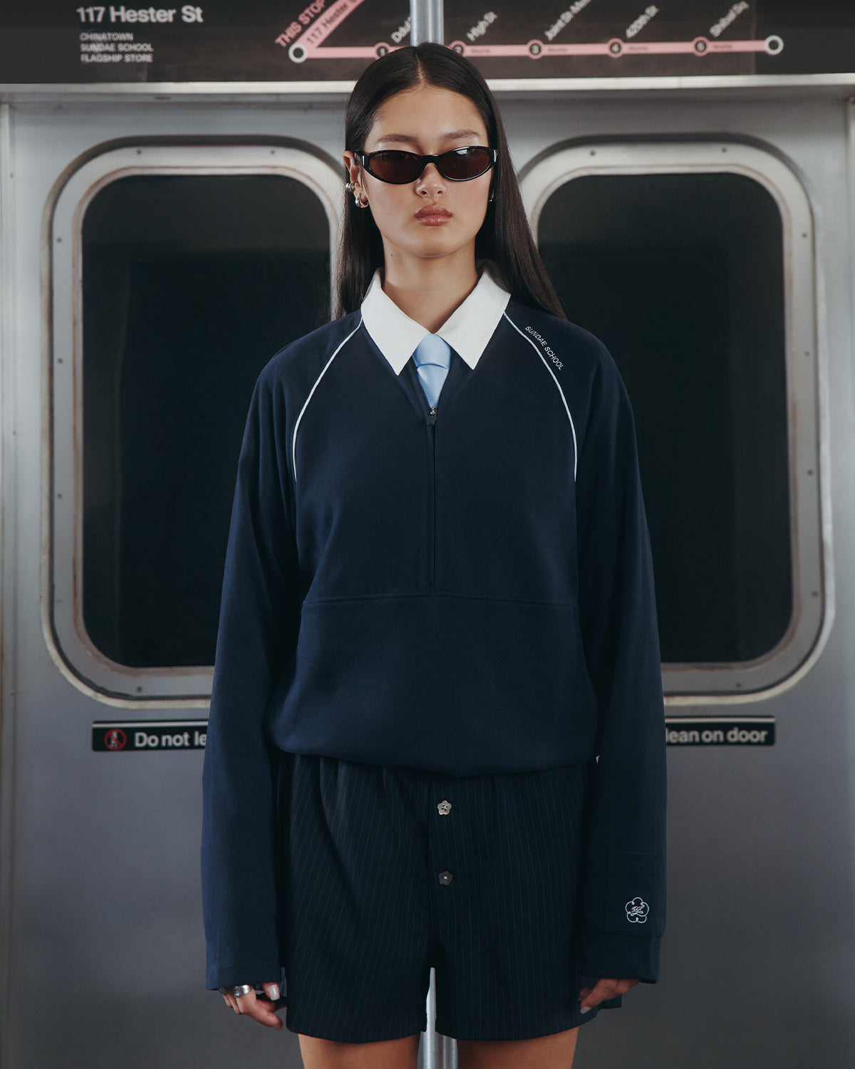 A woman with long dark hair and sunglasses wears the Sundae School NAVY OFFICE QUARTER ZIP, light blue tie, and dark shorts as she stands by subway doors beneath a map and a “117 Hester St” sign.