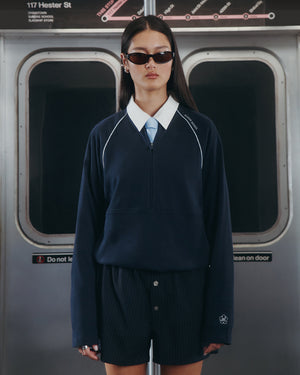 A woman with long dark hair and sunglasses wears the Sundae School NAVY OFFICE QUARTER ZIP, light blue tie, and dark shorts as she stands by subway doors beneath a map and a “117 Hester St” sign.