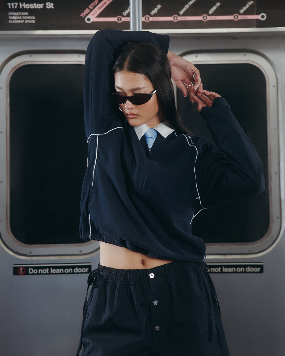 A person wearing the Sundae School NAVY OFFICE QUARTER ZIP and sunglasses poses with one arm raised inside a subway train, standing in front of closed doors with a "Do not lean on door" sign.