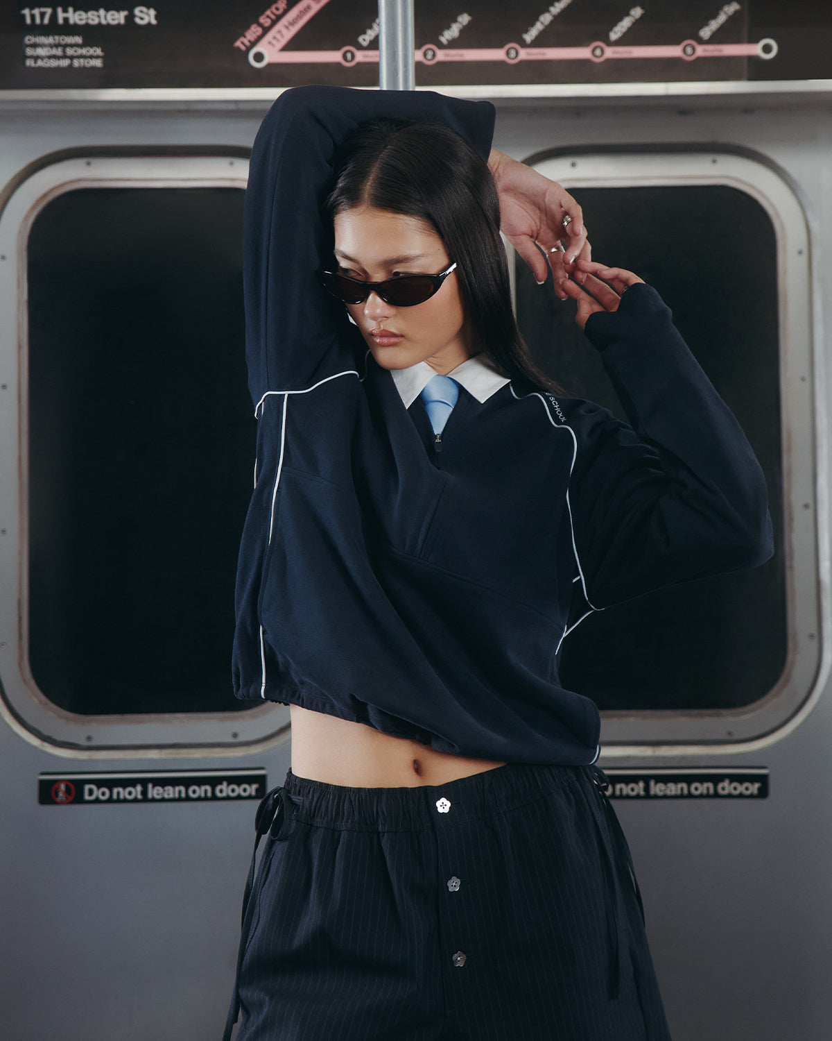 A person wearing the Sundae School NAVY OFFICE QUARTER ZIP and sunglasses poses with one arm raised inside a subway train, standing in front of closed doors with a "Do not lean on door" sign.