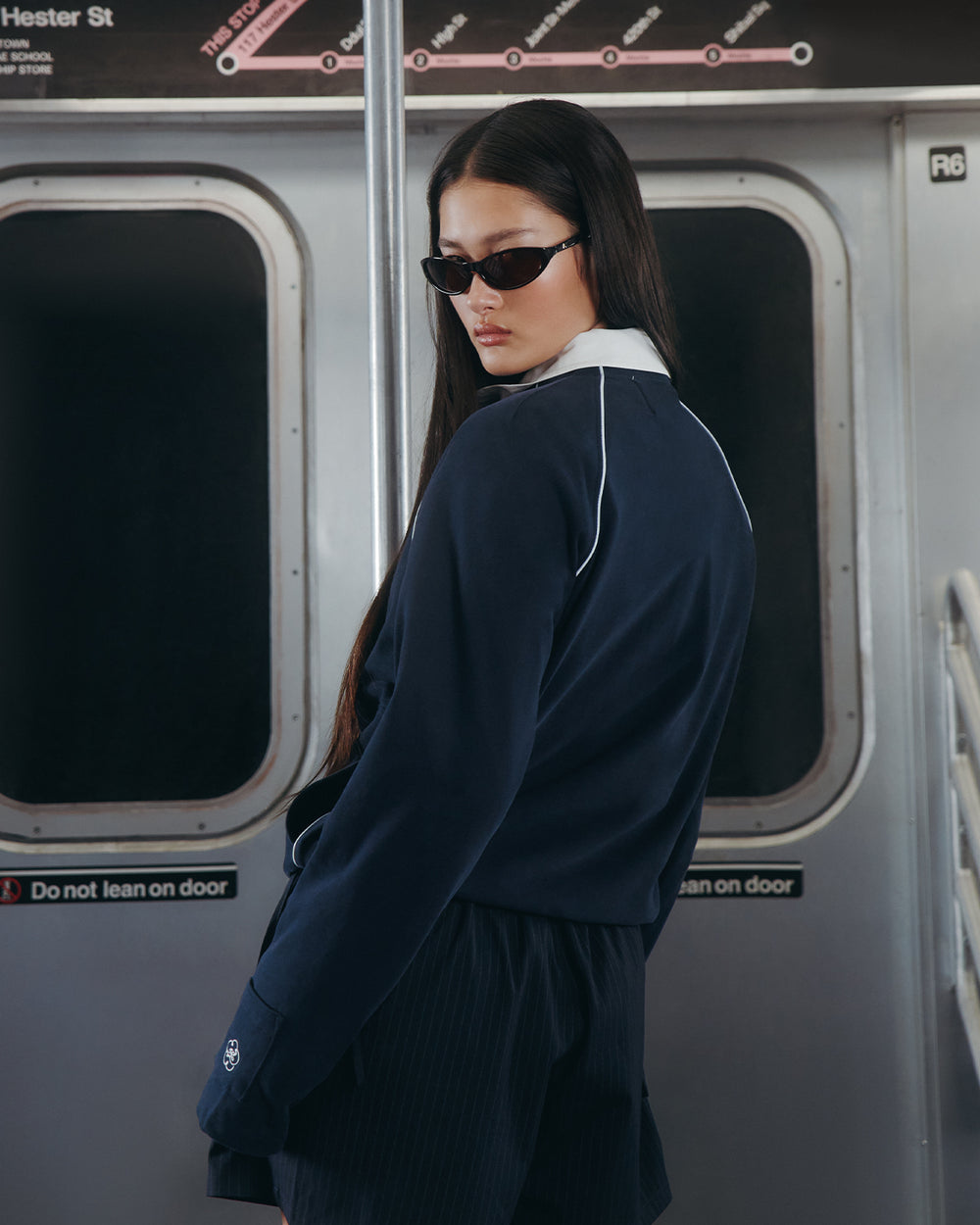 A woman with long dark hair, sunglasses, and shorts stands in a subway car by a pole, wearing the Sundae School NAVY OFFICE QUARTER ZIP. Subway door signs read “Do not lean on door.”.