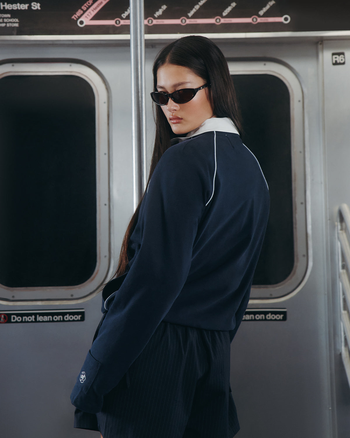 A woman with long dark hair, sunglasses, and shorts stands in a subway car by a pole, wearing the Sundae School NAVY OFFICE QUARTER ZIP. Subway door signs read “Do not lean on door.”.