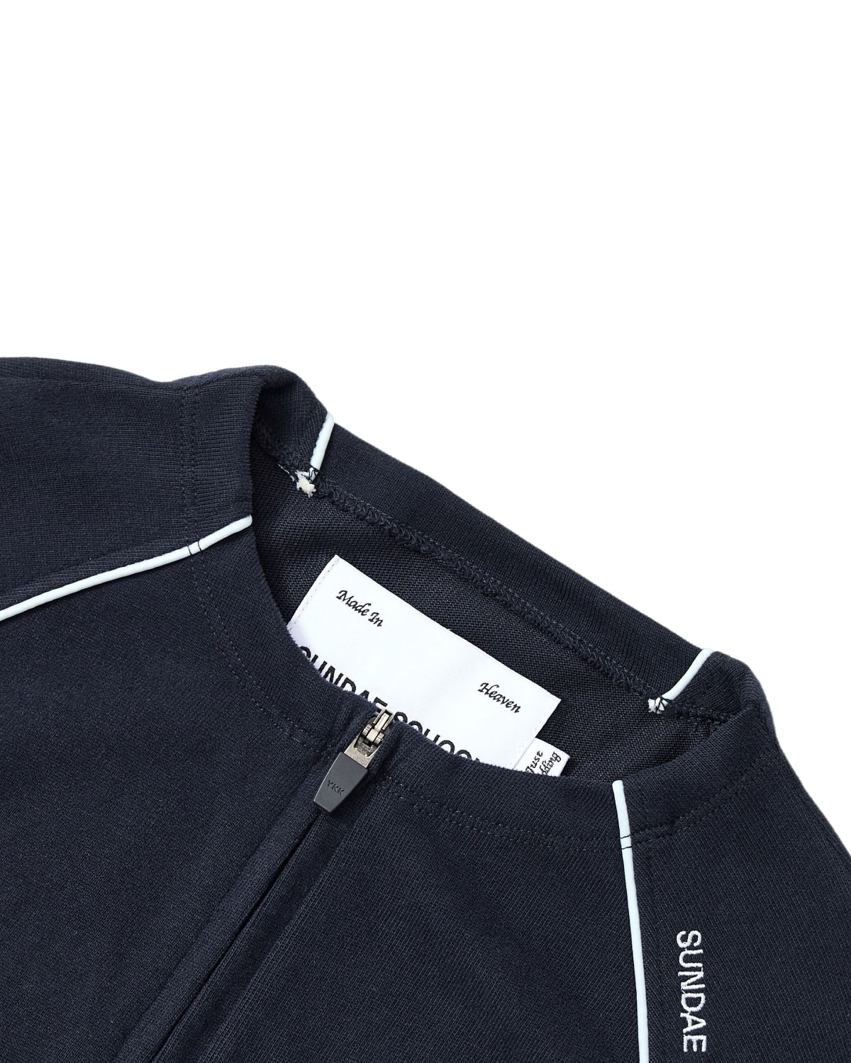 Close-up of the Sundae School NAVY OFFICE QUARTER ZIP, a navy blue cotton jersey zip-up featuring white piping and “SUNDAE” text on the collar. A white clothing label is visible inside near the zipper.