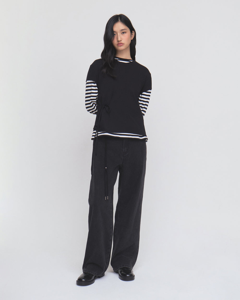 A woman stands against a plain white background, wearing the BLACK ORIGAMI GORUM TEE by Sundae School, which features black-and-white striped sleeves. She pairs the top with loose black pants and black shoes. Her long dark hair falls loosely, and she has a neutral expression.