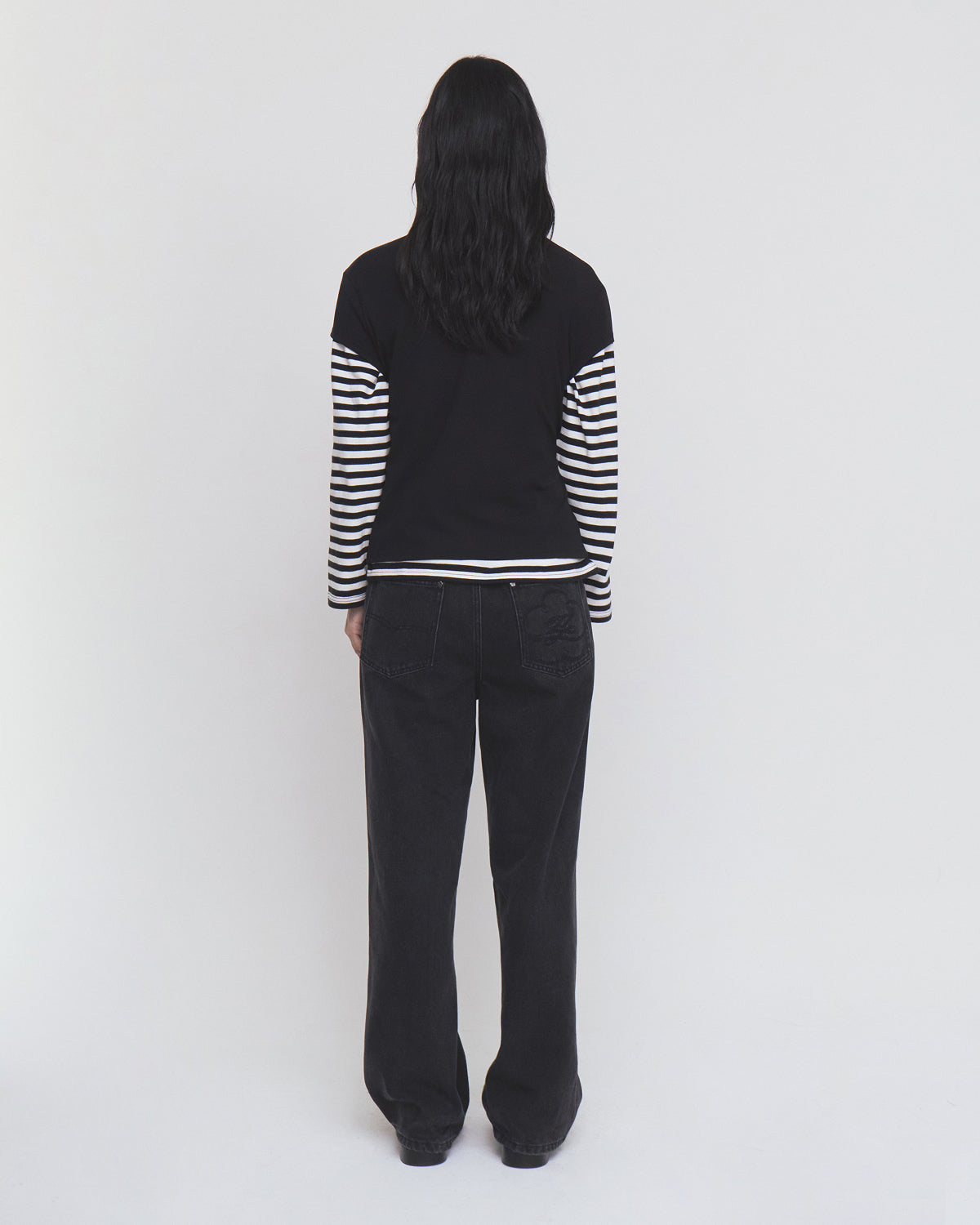 A person with long dark hair stands facing away against a plain light background, wearing Sundae School’s BLACK ORIGAMI GORUM TEE, which features black and white striped sleeves, paired with loose black pants.