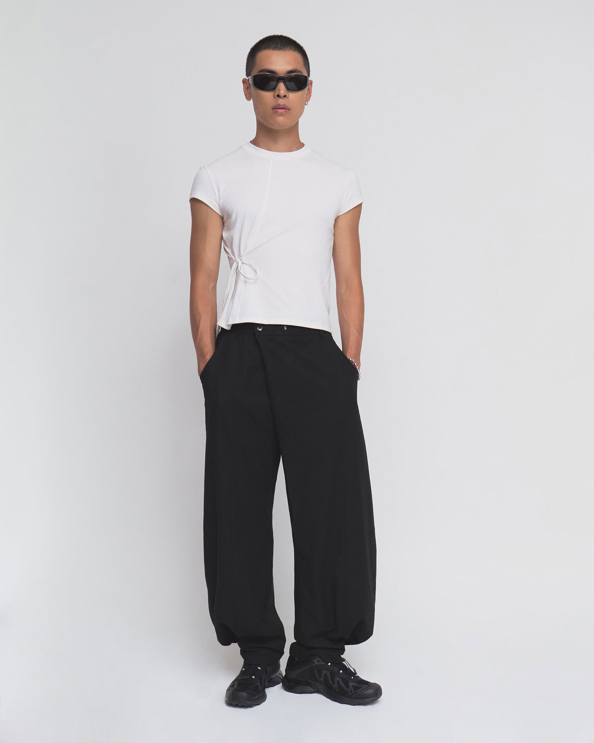 A person wearing sunglasses, the Sundae School IVORY ORIGAMI GORUM TEE with a side tie detail, loose black pants, and black sneakers stands against a plain white background.