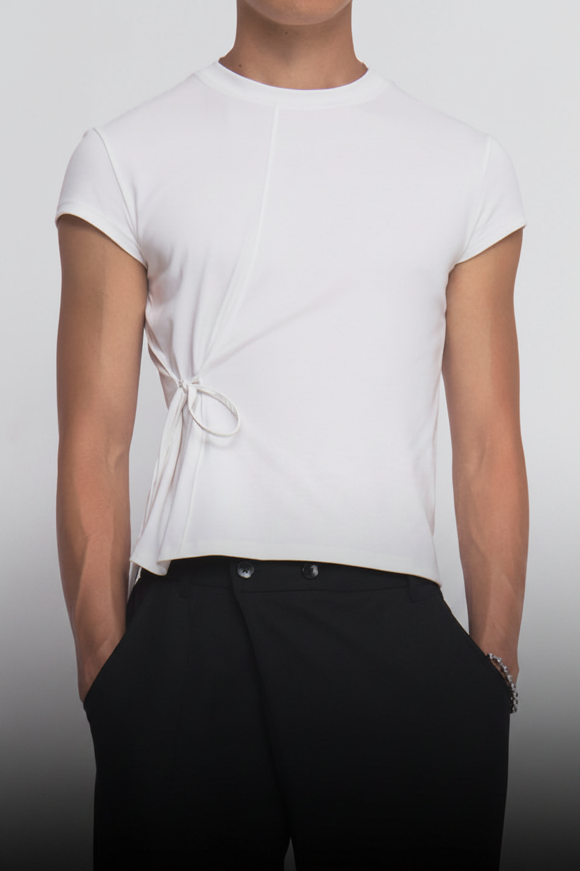 A person wearing sunglasses, the Sundae School IVORY ORIGAMI GORUM TEE with a side tie detail, loose black pants, and black sneakers stands against a plain white background.