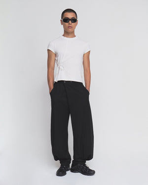 A person wearing sunglasses, the Sundae School IVORY ORIGAMI GORUM TEE with a side tie detail, loose black pants, and black sneakers stands against a plain white background.