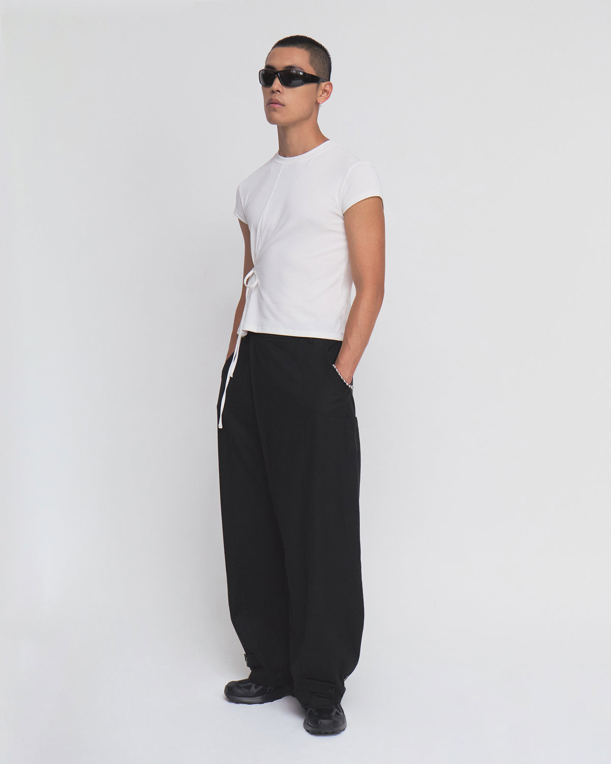 A person wearing the Sundae School IVORY ORIGAMI GORUM TEE—a fitted white t-shirt with side ties—paired with loose black pants, black sunglasses, and black shoes stands against a plain white background with hands in their pockets.