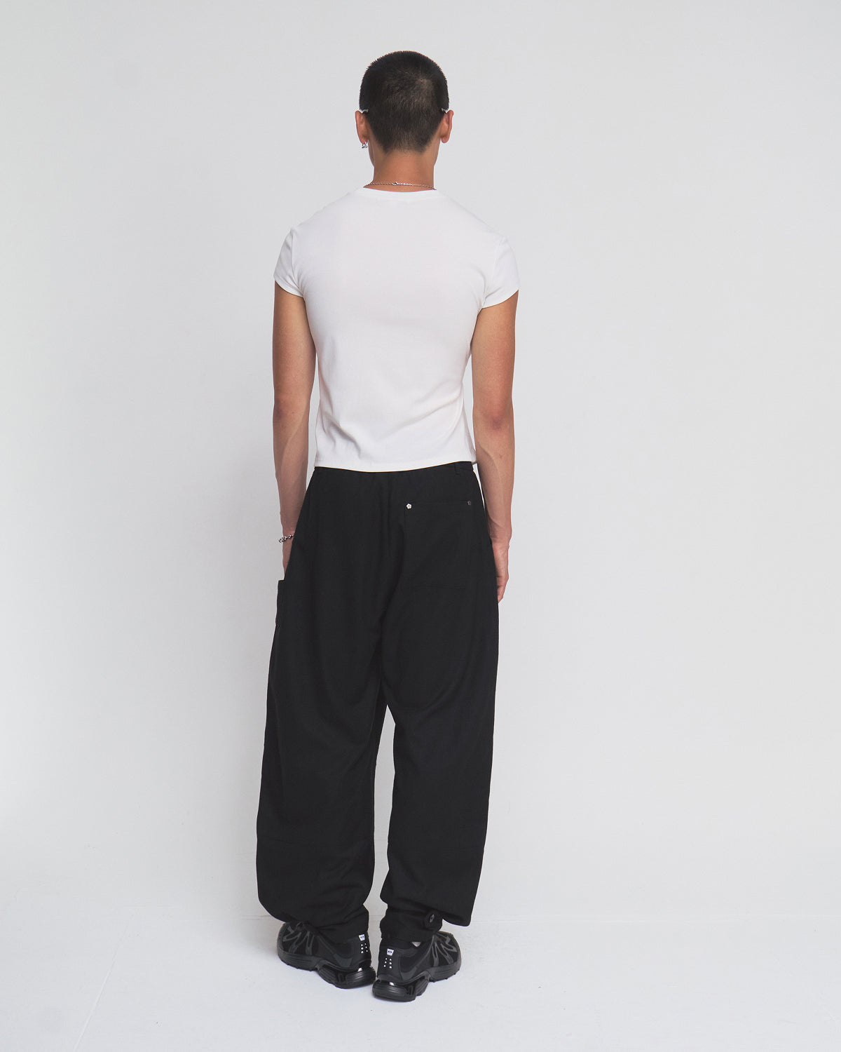 A person with short hair stands facing away, wearing the Sundae School IVORY ORIGAMI GORUM TEE, loose black pants, and black shoes against a plain white background.