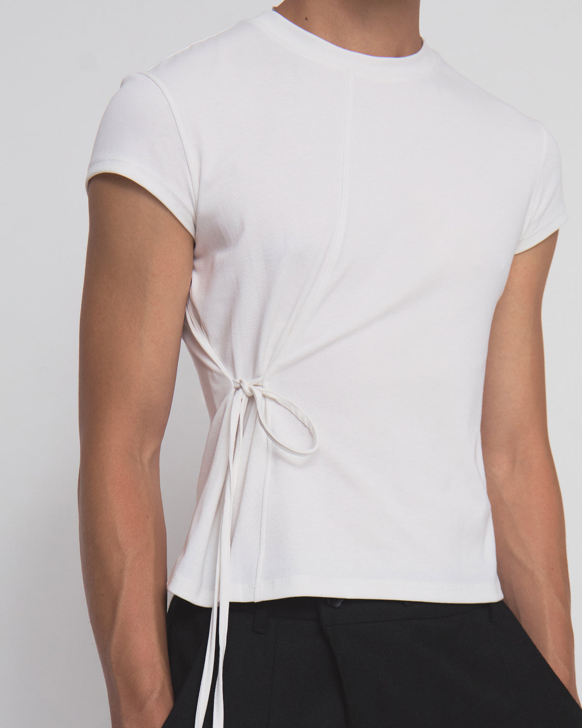 A person wearing the Sundae School IVORY ORIGAMI GORUM TEE, a fitted white short-sleeve top with side tie detail, paired with black pants and standing against a plain light background.