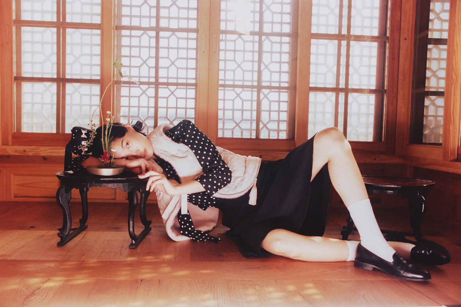 A woman reclines on a wooden floor in a traditional room with large window panes, wearing Sundae School Pink Silver Baeja Vest.