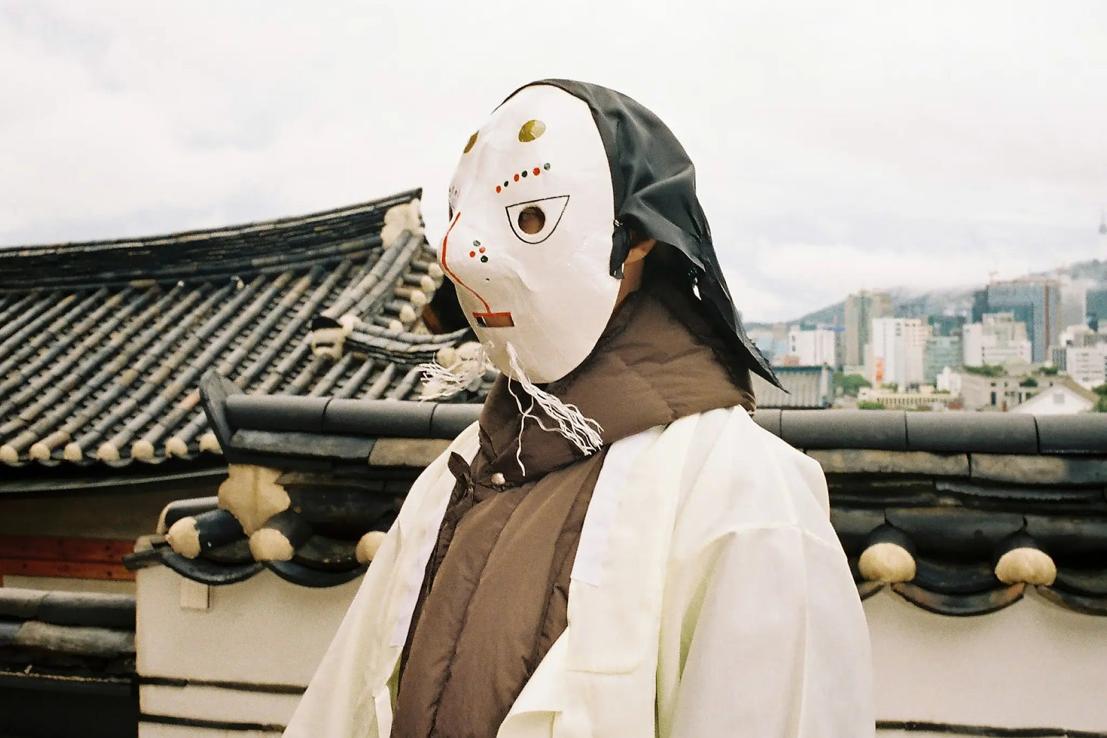 A person wearing a Korean traditional white and black mask with Sundae School Heavyweight Mochi Puffer, standing outdoors in front of a traditional tiled roof and a Seoul skyline under an overcast sky.