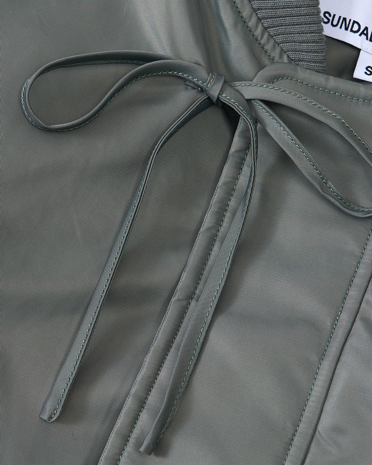 Close-up of the KHAKI FLOWER CHANNELED BOMBER in sage green by Sundae School, featuring a tied front bow, visible stitching details, and a partially visible clothing label showing the brand and size S at the top right corner.