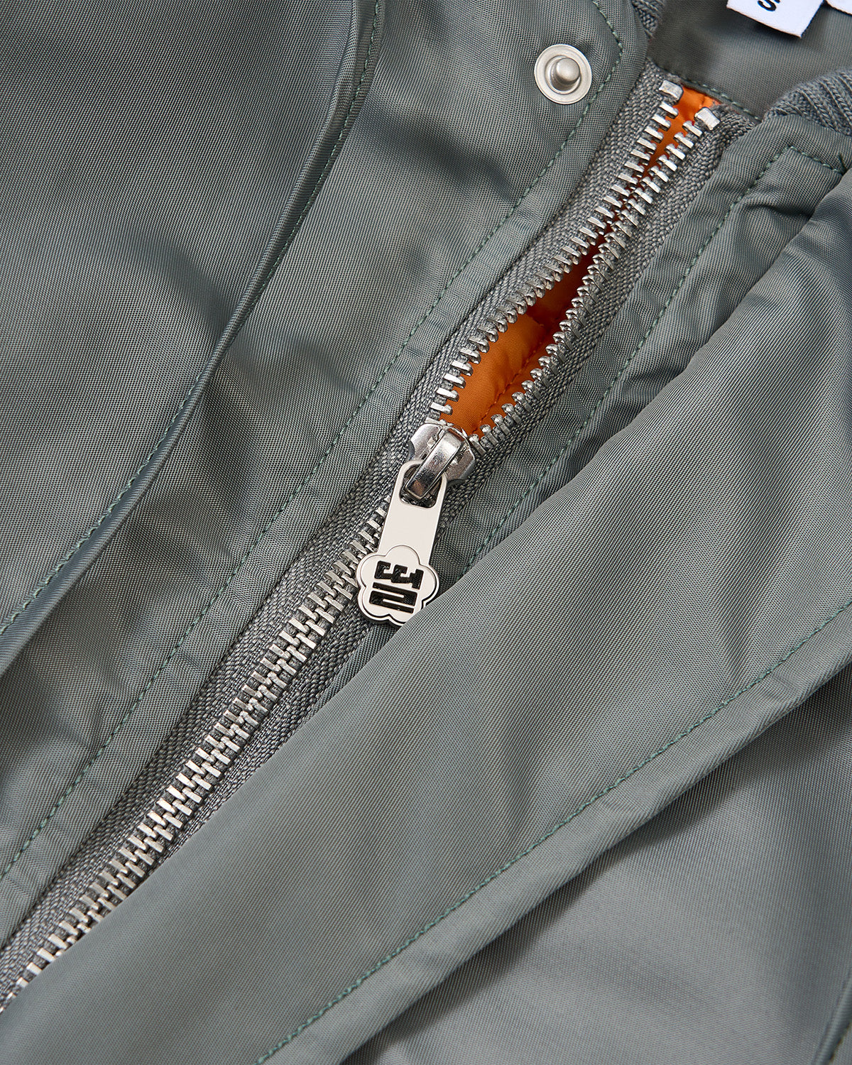 Close-up of the silver zipper on Sundae School's KHAKI FLOWER CHANNELED BOMBER, revealing a hint of bright orange lining beneath the olive green exterior.