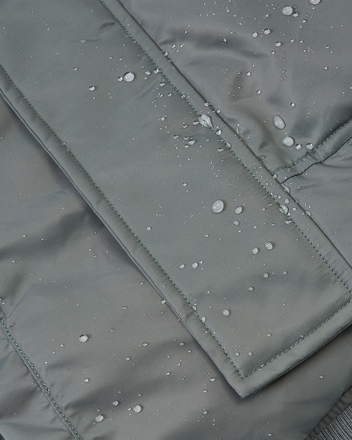 Close-up of water droplets beading on the gray, water-resistant surface of the Sundae School KHAKI FLOWER CHANNELED BOMBER, highlighting its waterproof qualities.