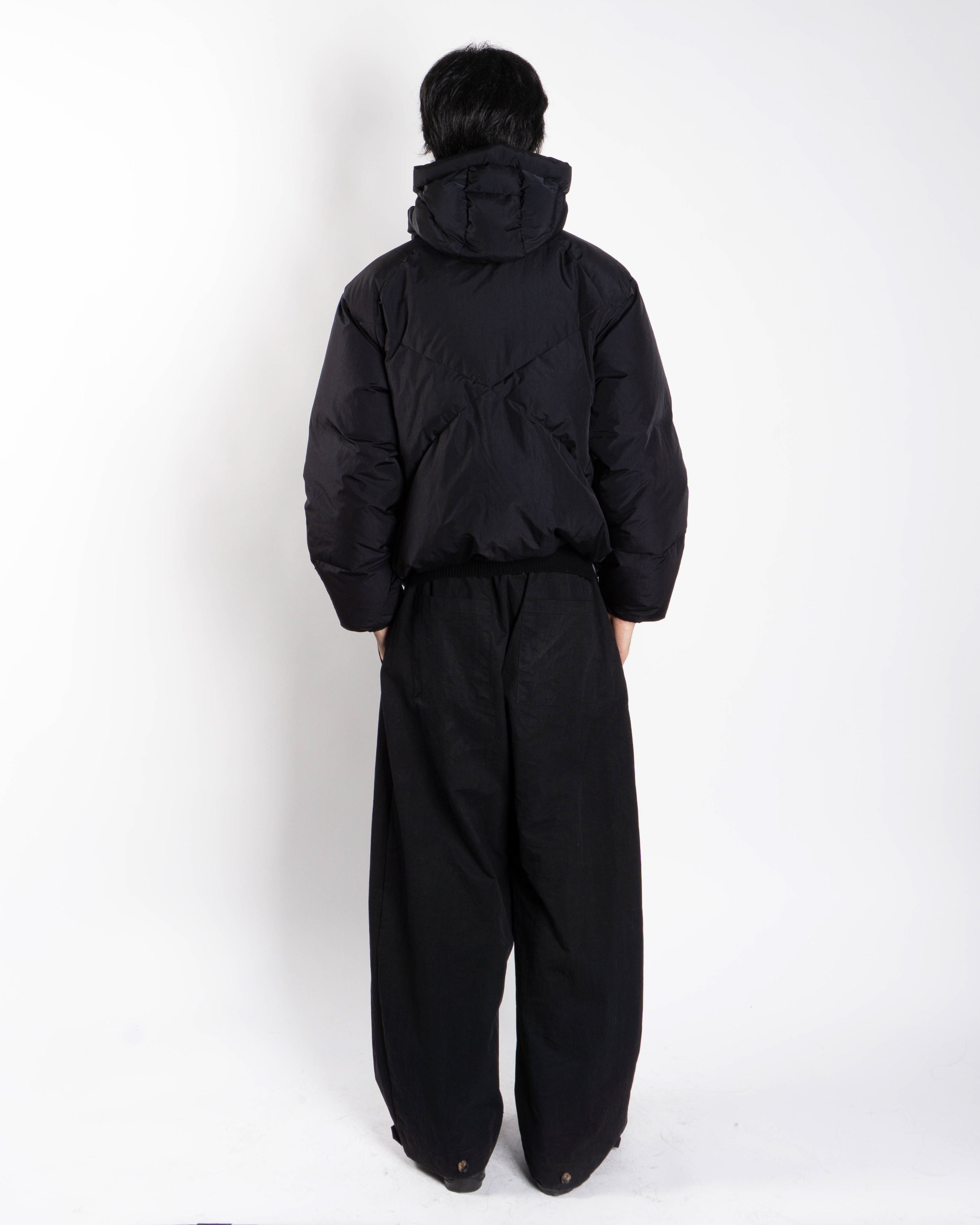 A person faces away from the camera, wearing Sundae School's BLACK SKY BLUE ORIGAMI PUFFER and loose black pants against a plain white background.