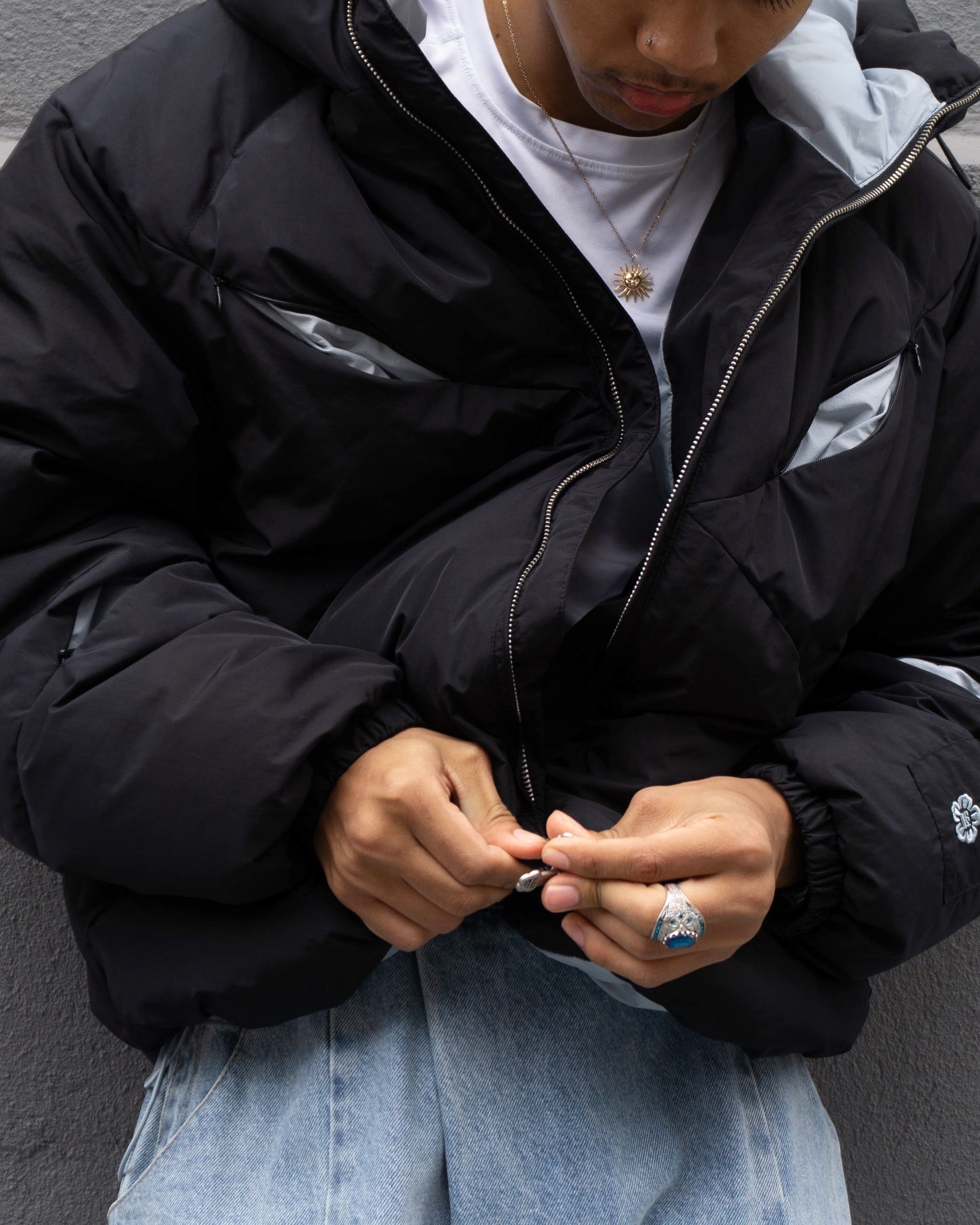 A person wears the Sundae School BLACK SKY BLUE ORIGAMI PUFFER over a white shirt and zips it up. Light blue jeans, a sun-shaped pendant necklace, and a ring complete the look. Their face is partially out of frame.