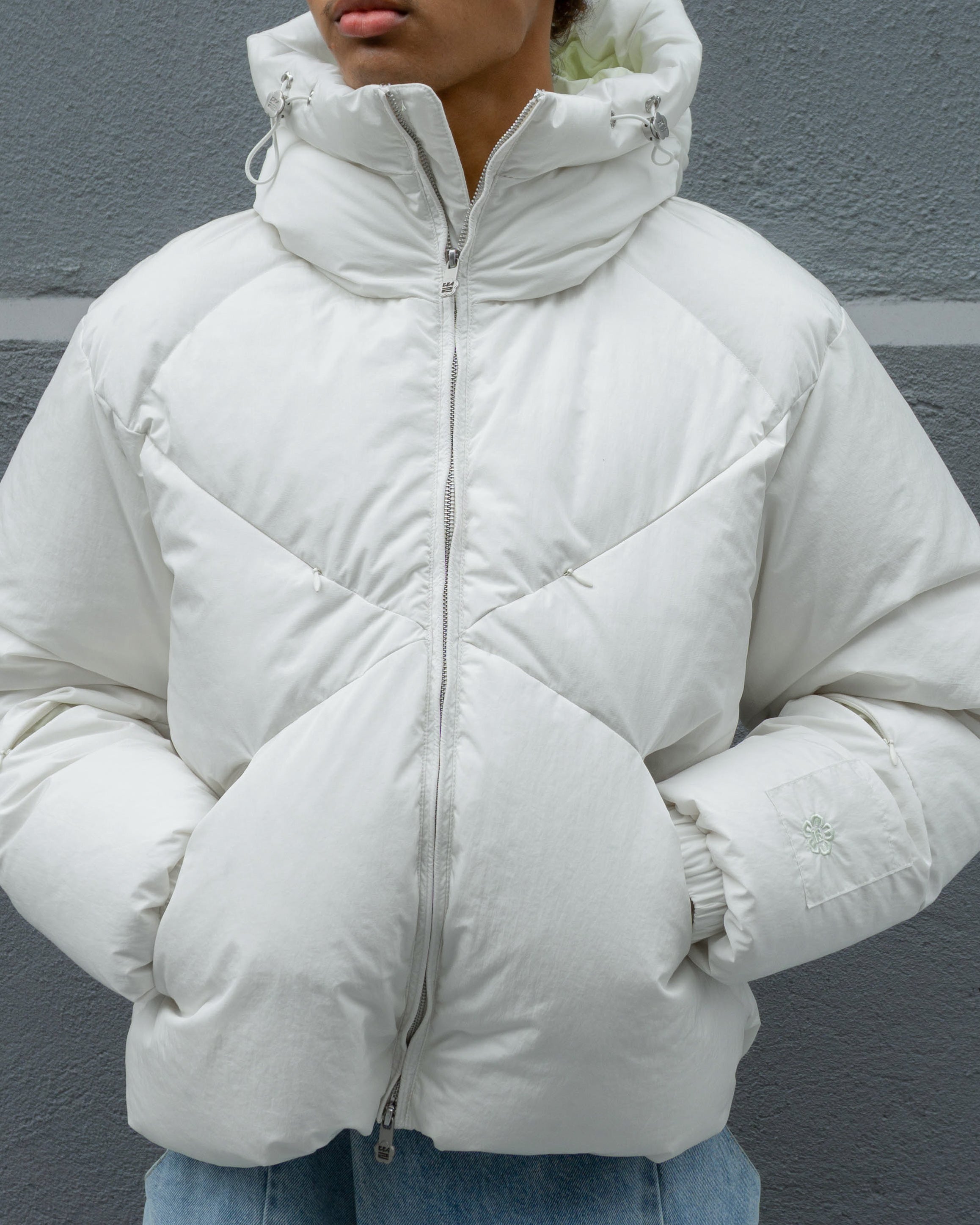 A person stands against a gray wall wearing the Sundae School IVORY LIME ORIGAMI PUFFER, a white hooded puffer jacket with a quilted pattern and silver zipper, hands in pockets, face partially out of frame.