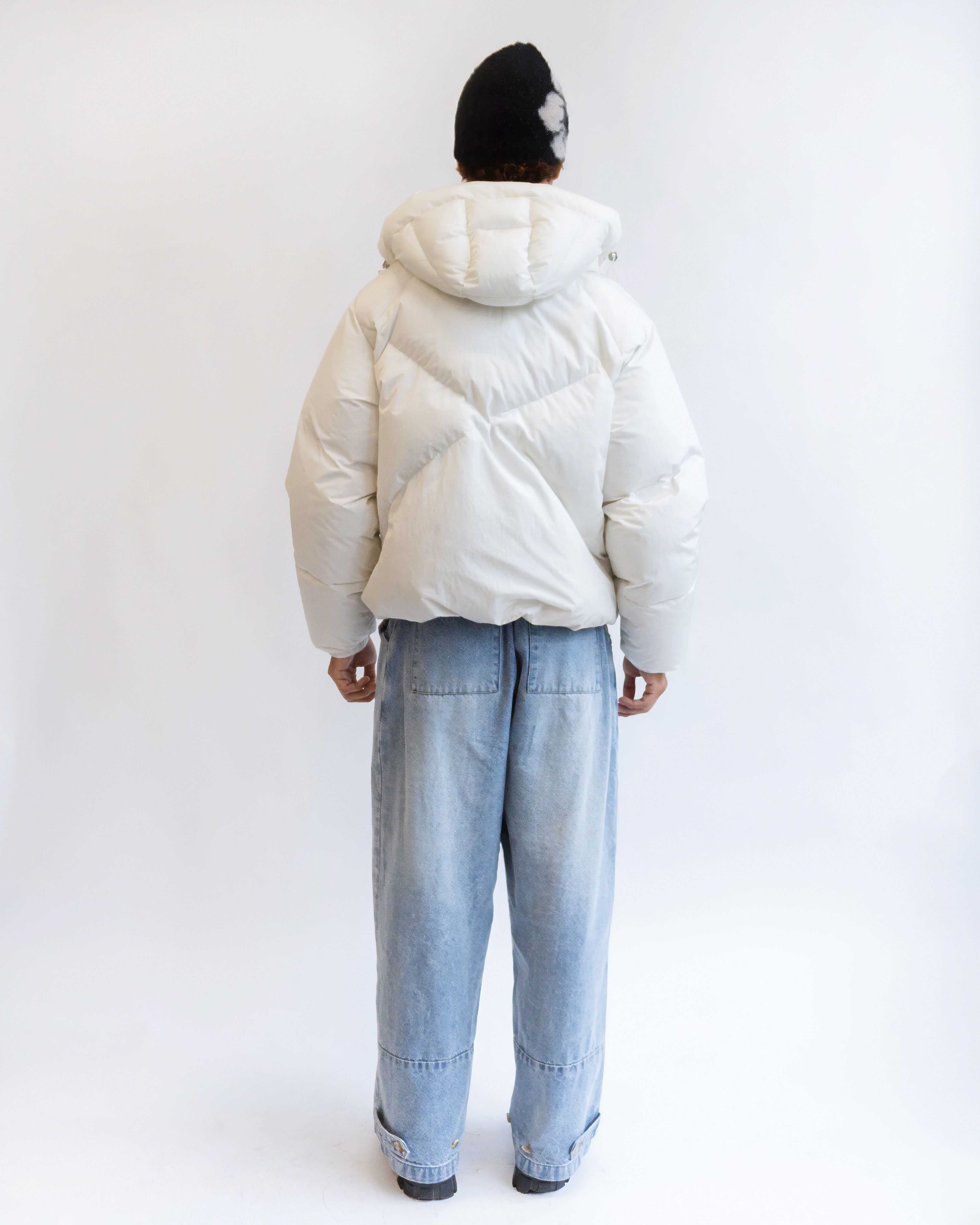 A person stands with their back to the camera, wearing Sundae School’s IVORY LIME ORIGAMI PUFFER, loose light blue jeans, and a black beanie with a white pattern, set against a plain white background.