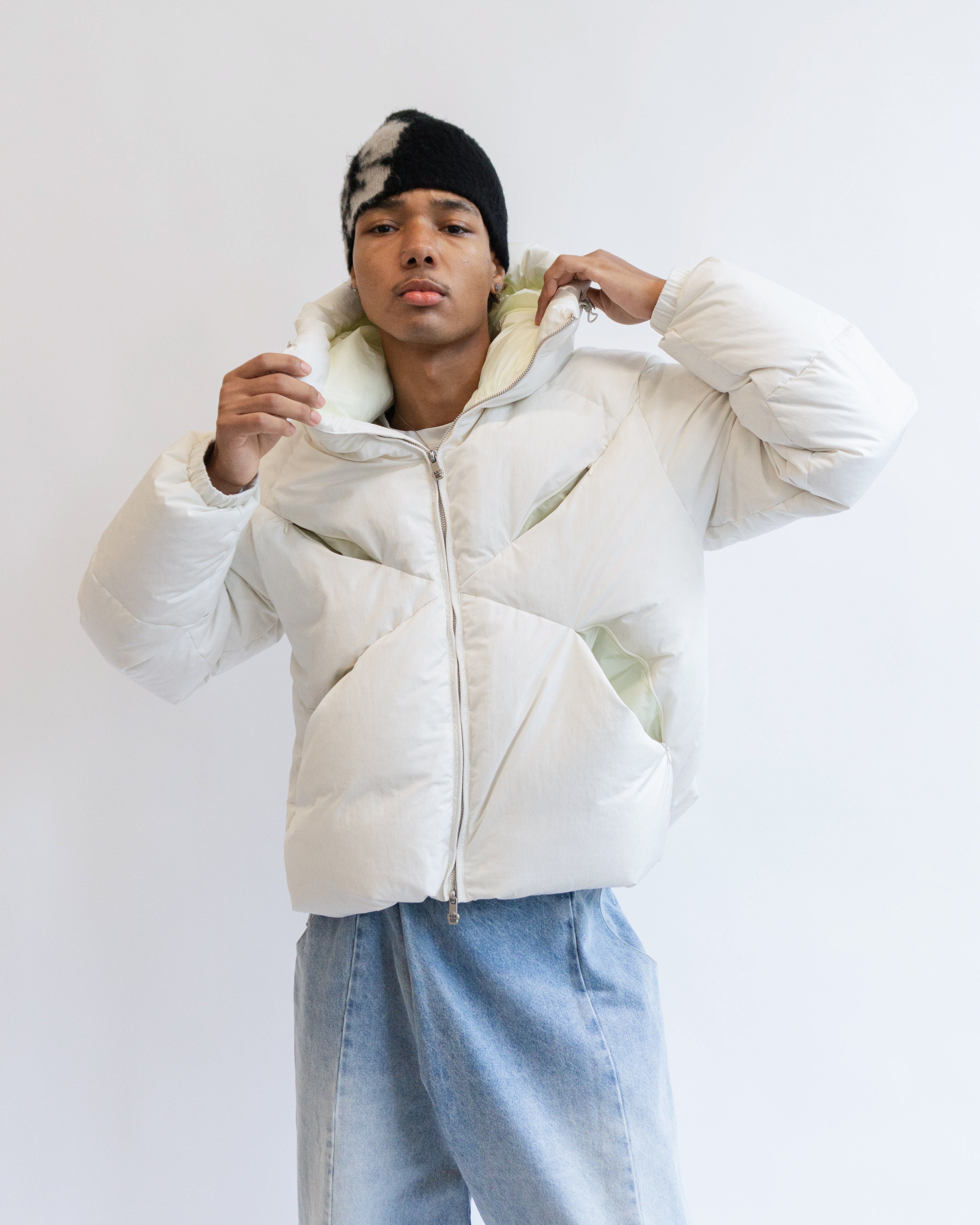 A person wearing the Sundae School IVORY LIME ORIGAMI PUFFER and light blue jeans adjusts the hood while facing the camera. They also wear a black and white knit hat, standing against a plain white background.