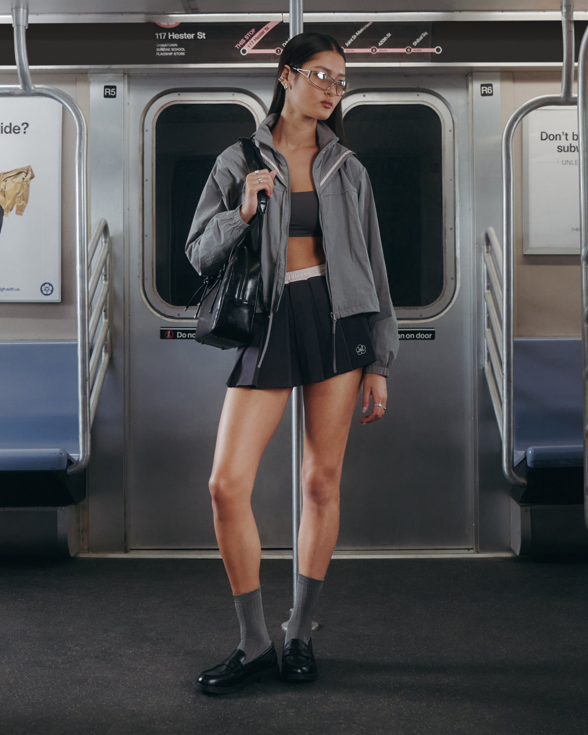 A person in the Sundae School CHARCOAL SAILOR WINDBREAKER, crop top, dark shorts, gray socks, and black shoes stands inside a mostly empty subway train holding a black bag.