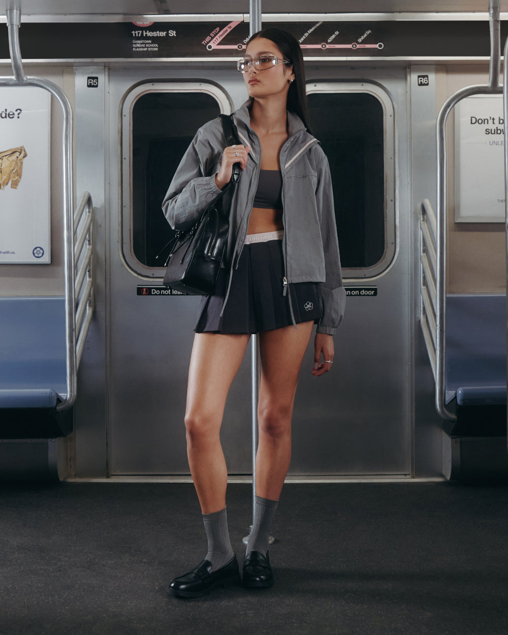 A woman wearing Sundae School's CHARCOAL PLEATED JERSEY MINI SKIRT, a gray jacket, and crop top stands in an empty subway car, holding a black handbag and gazing to the side.