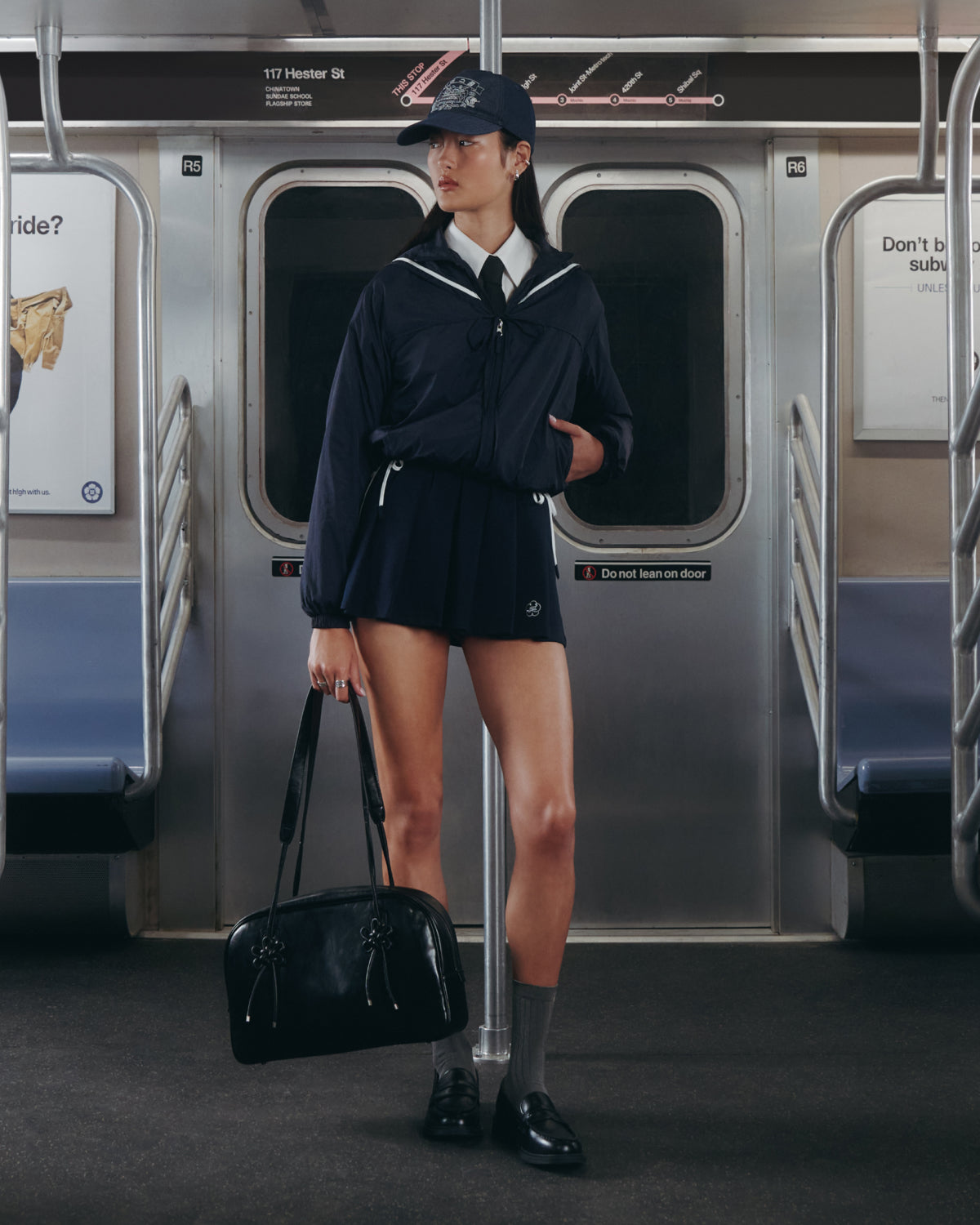 In a subway car, a person stands holding a black bag, dressed in Sundae School's NAVY PLEATED JERSEY MINI SKIRT with matching sailor-style accents; empty seats and ads can be seen in the background.