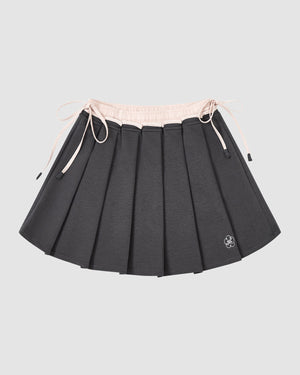 The CHARCOAL PLEATED JERSEY MINI SKIRT by Sundae School features dark gray knife pleats, a light pink elastic waistband with drawstrings, and a small white embroidered flower at the hem, shown against a plain light gray background.