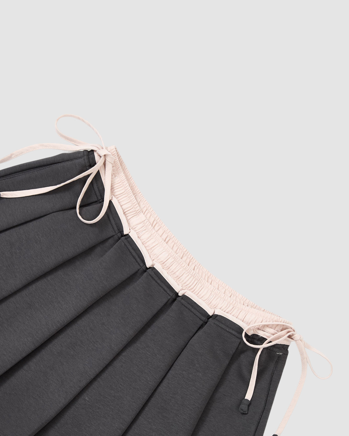 A close-up of the CHARCOAL PLEATED JERSEY MINI SKIRT by Sundae School, featuring knife pleats, a light pink elastic waistband with drawstrings, shown against a plain light background.