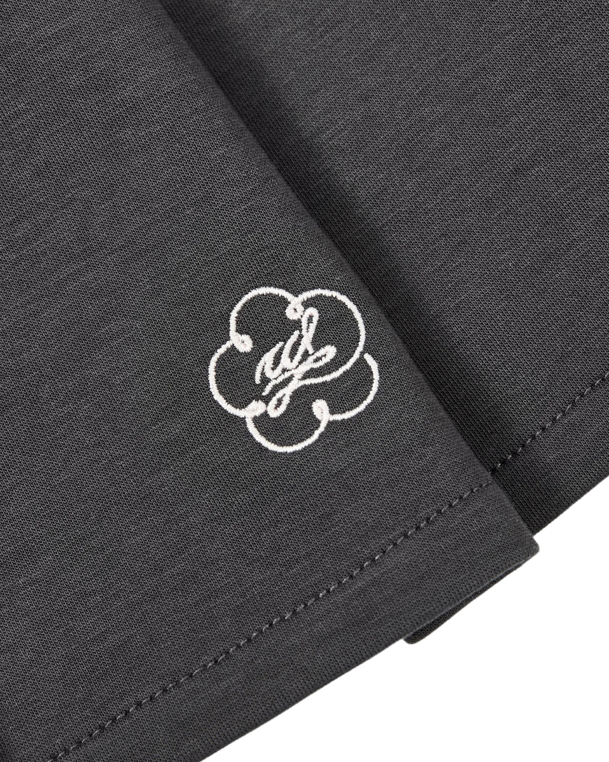A close-up of the corner of the CHARCOAL PLEATED JERSEY MINI SKIRT by Sundae School, made from a dark gray cotton-blend jersey, featuring white stitching and an embroidered white cloud-like logo with cursive text.